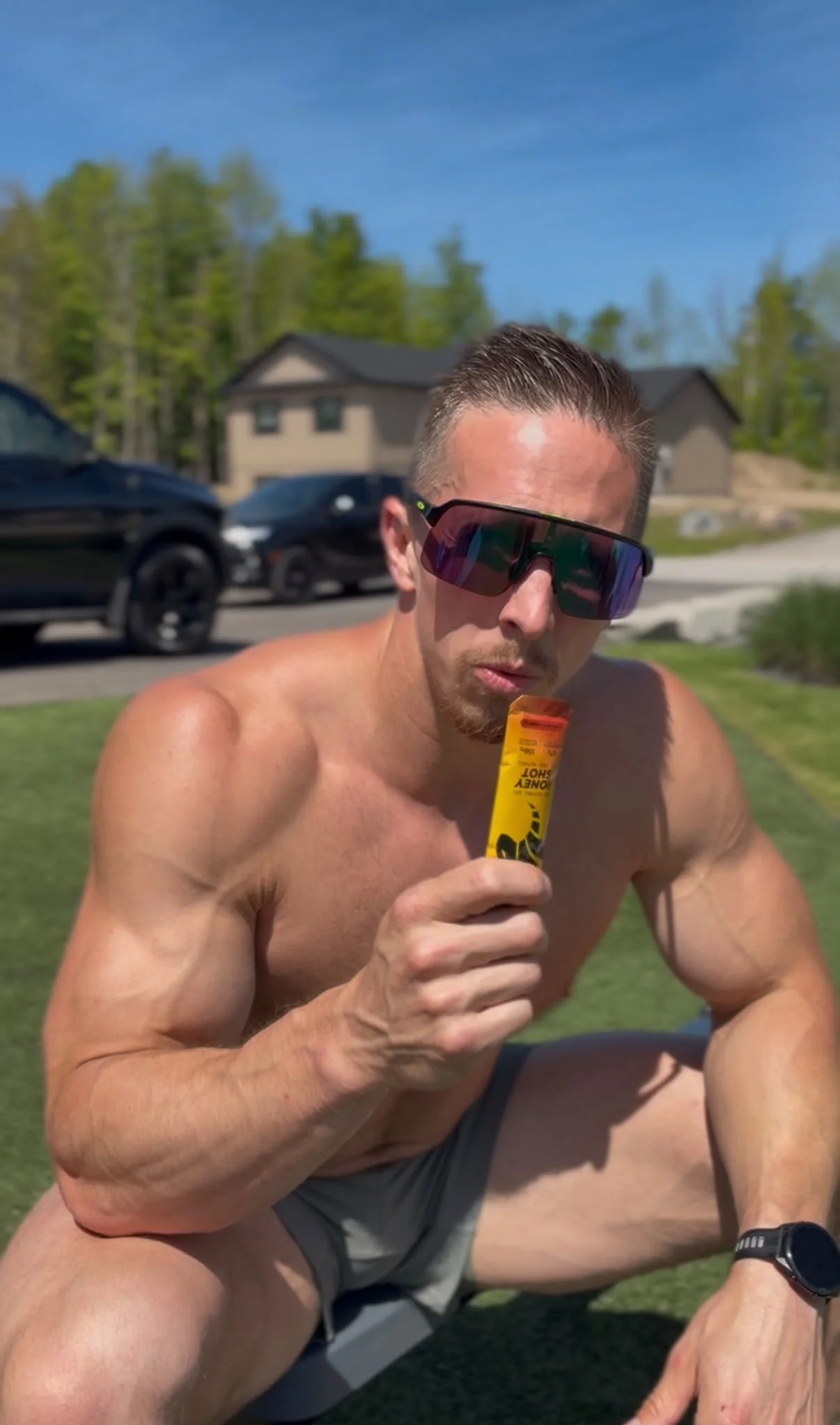 A shirtless man wearing sunglasses, holding a yellow sunscreen stick, sitting outdoors on grass with a house and parked cars in the background on a sunny day.