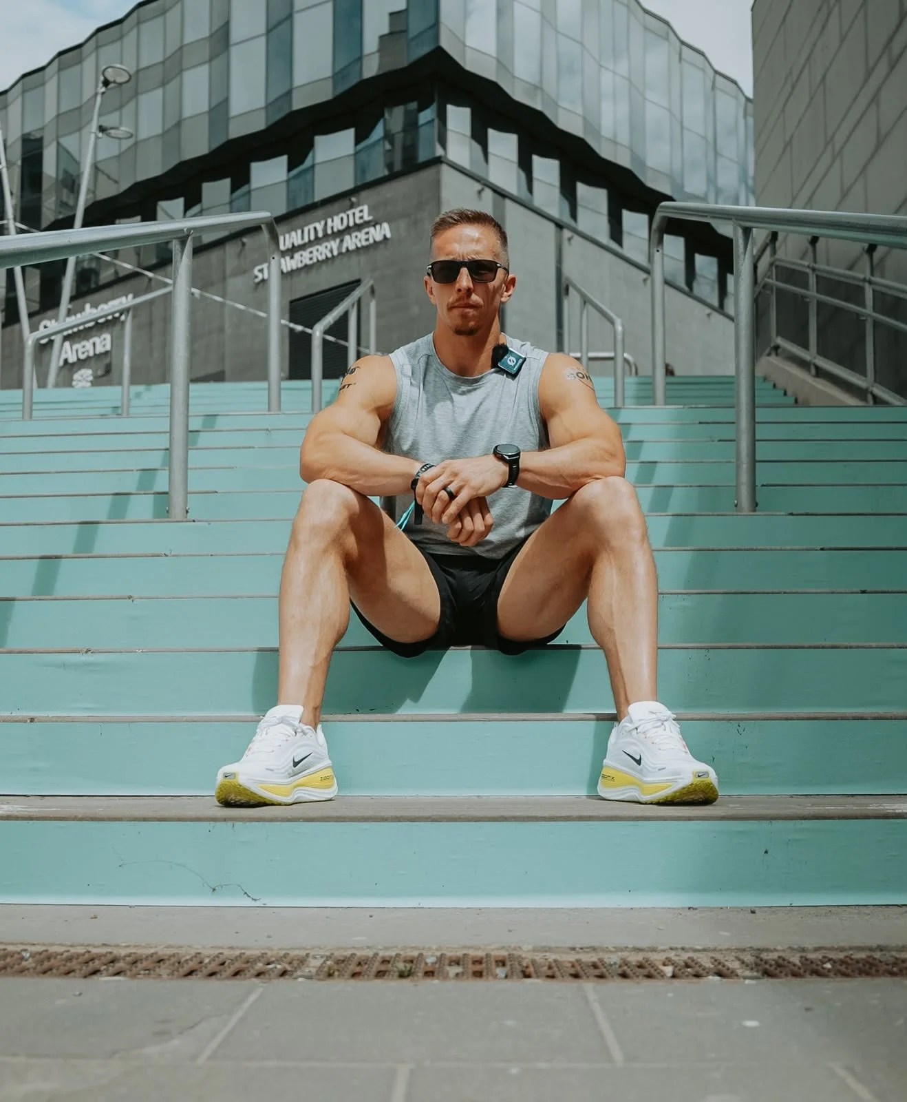 A man wearing sunglasses, a gray sleeveless shirt, black shorts, and white running shoes sitting on teal stairs outdoors in an urban setting with modern glass buildings in the background.