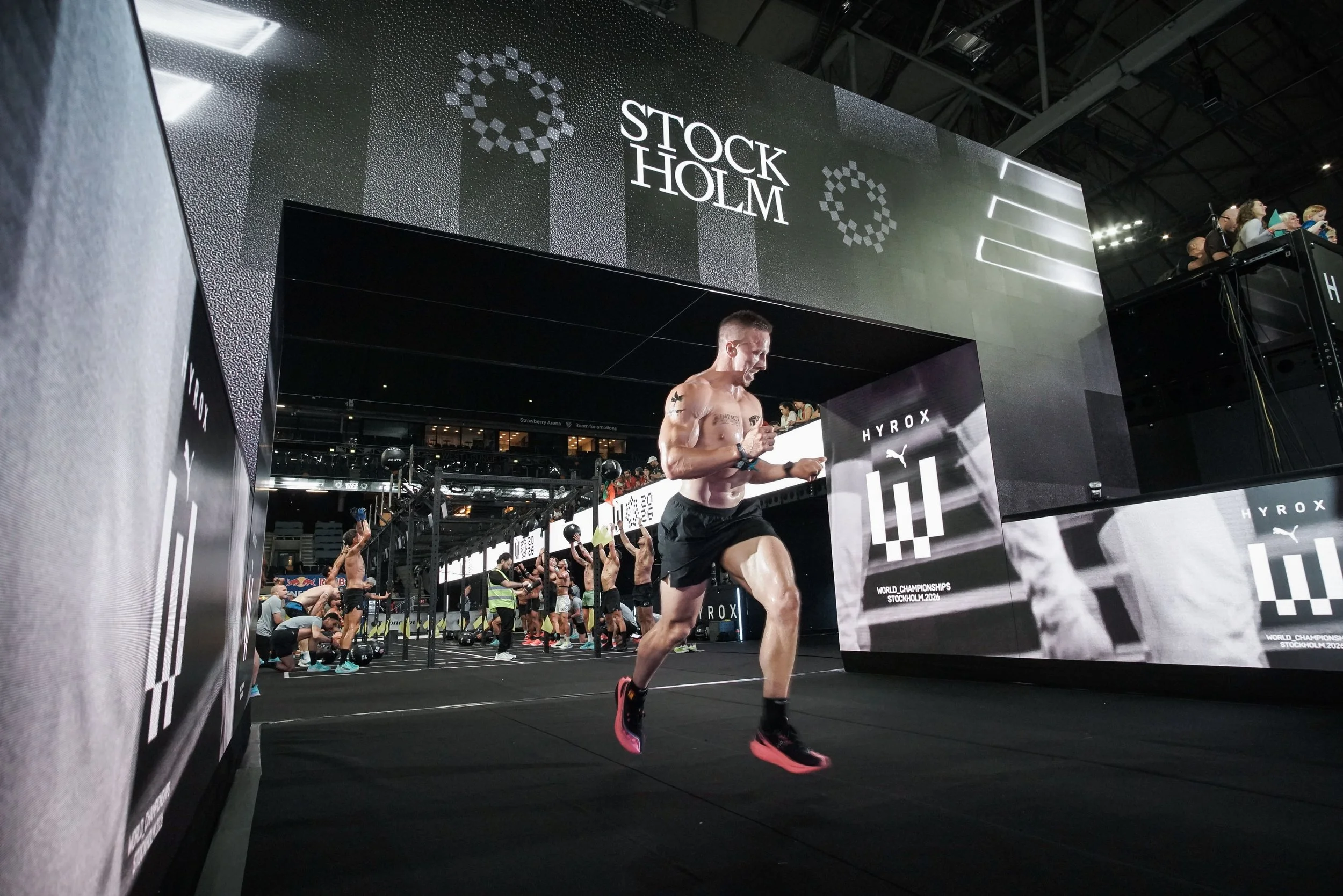 A muscular man running on a treadmill during a fitness event at Stockholm 2024 World Championships, with a large black and white sign above him reading 'Stockholm'.