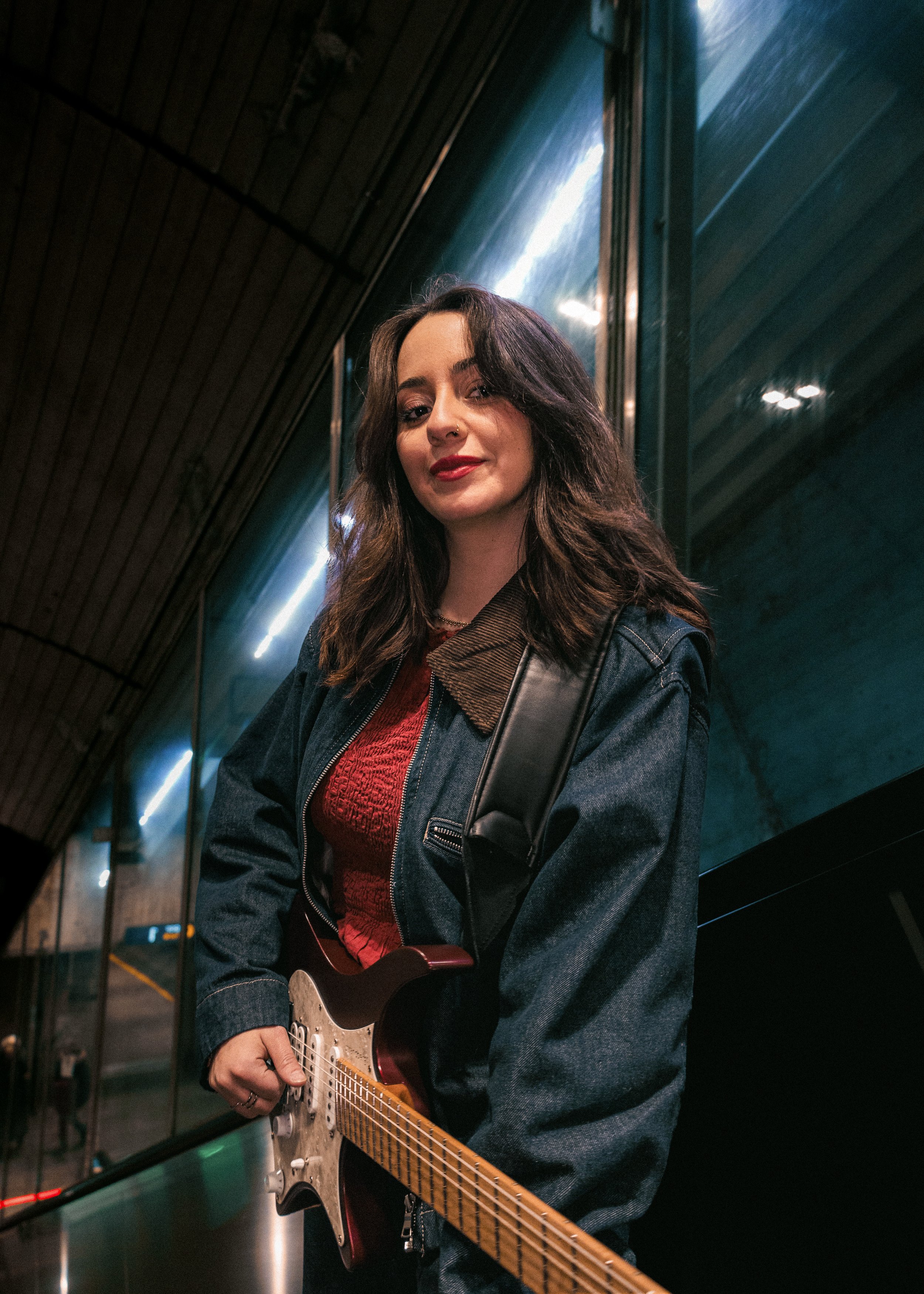 A woman with long brown hair holding an electric guitar, standing in a dimly lit, modern indoor space with wooden and reflective surfaces.