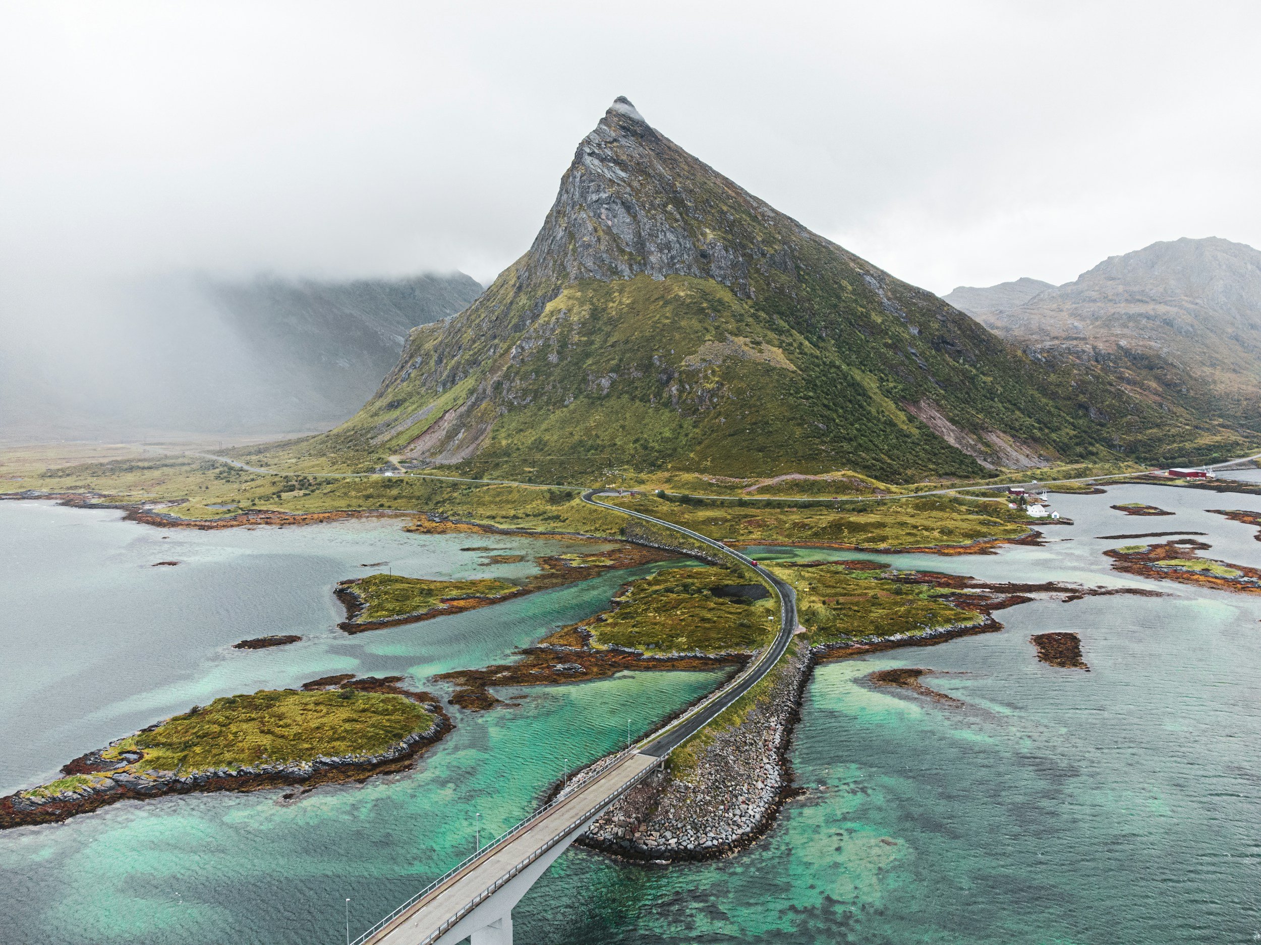 A bridge crossing over turquoise and dark blue water, leading towards a mountain with a sharp peak, surrounded by green vegetation and misty clouds.