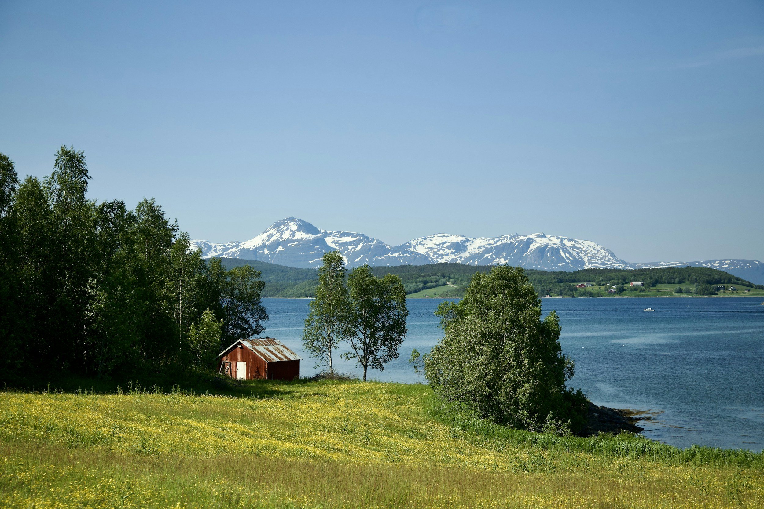 Scenic view of snow-capped mountains over a lake, with green trees and a small red barn in the foreground on a clear day.