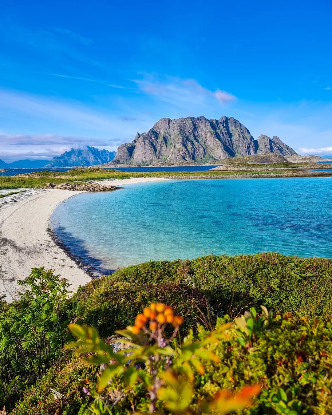 Scenic view of a beach with clear blue water, green vegetation, and a large mountain in the background under a partly cloudy sky.
