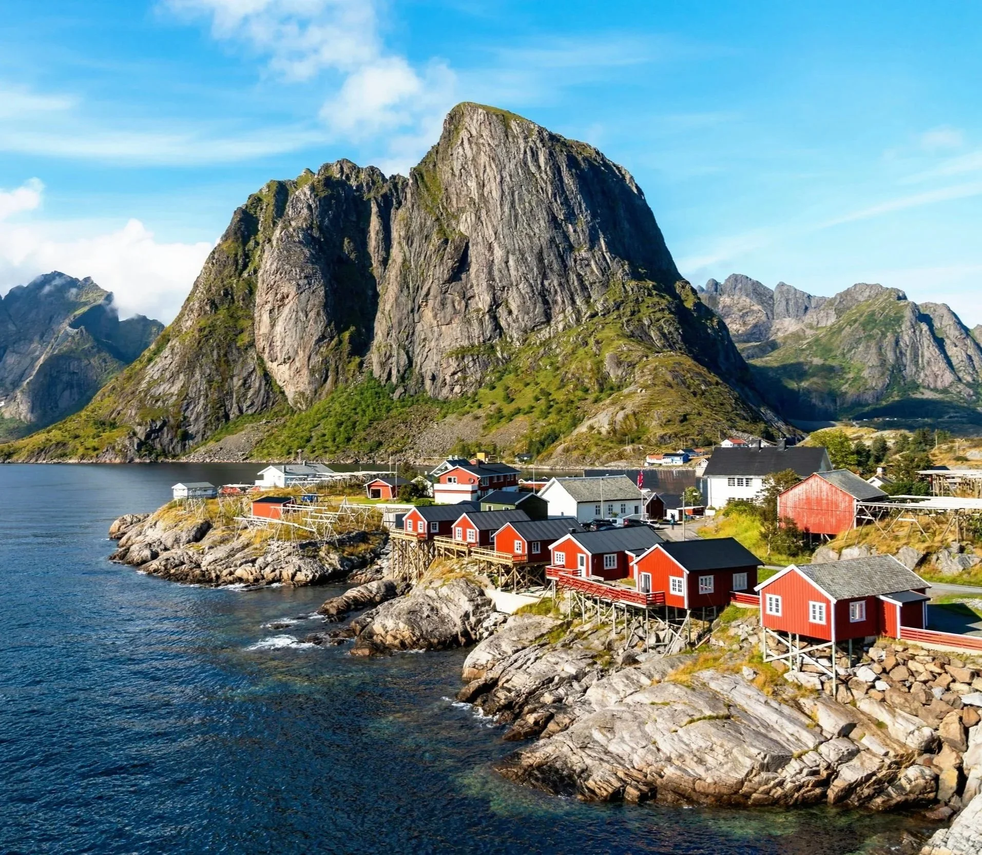 Colorful red, white, and black houses by the water with a large mountain in the background, under a blue sky with some clouds.