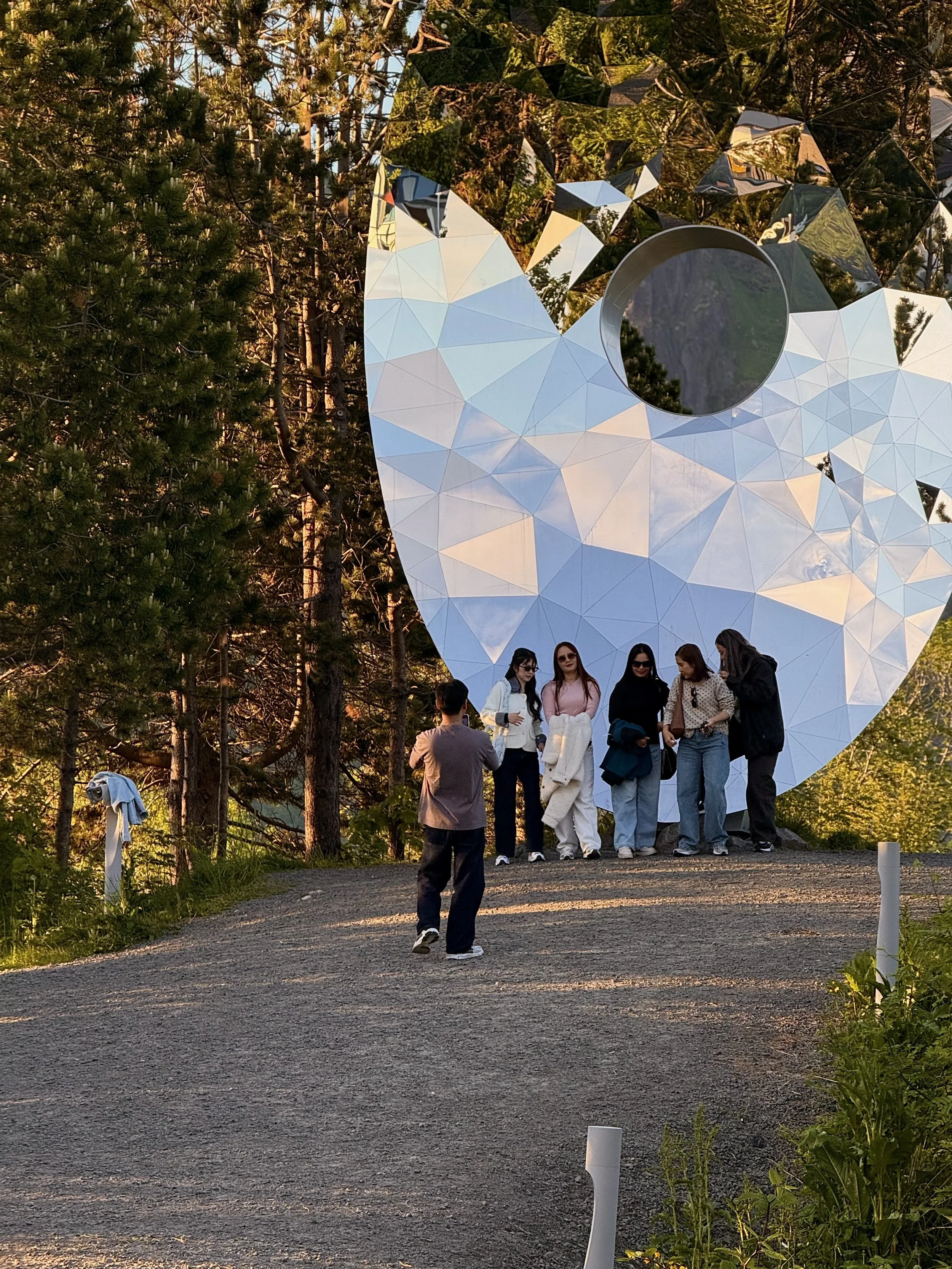 A group of six women standing on a gravel path in front of a large reflective sculpture that resembles a skull, surrounded by tall green trees. One man stands facing them, taking a photograph. The scene appears to be during the late afternoon or early evening.