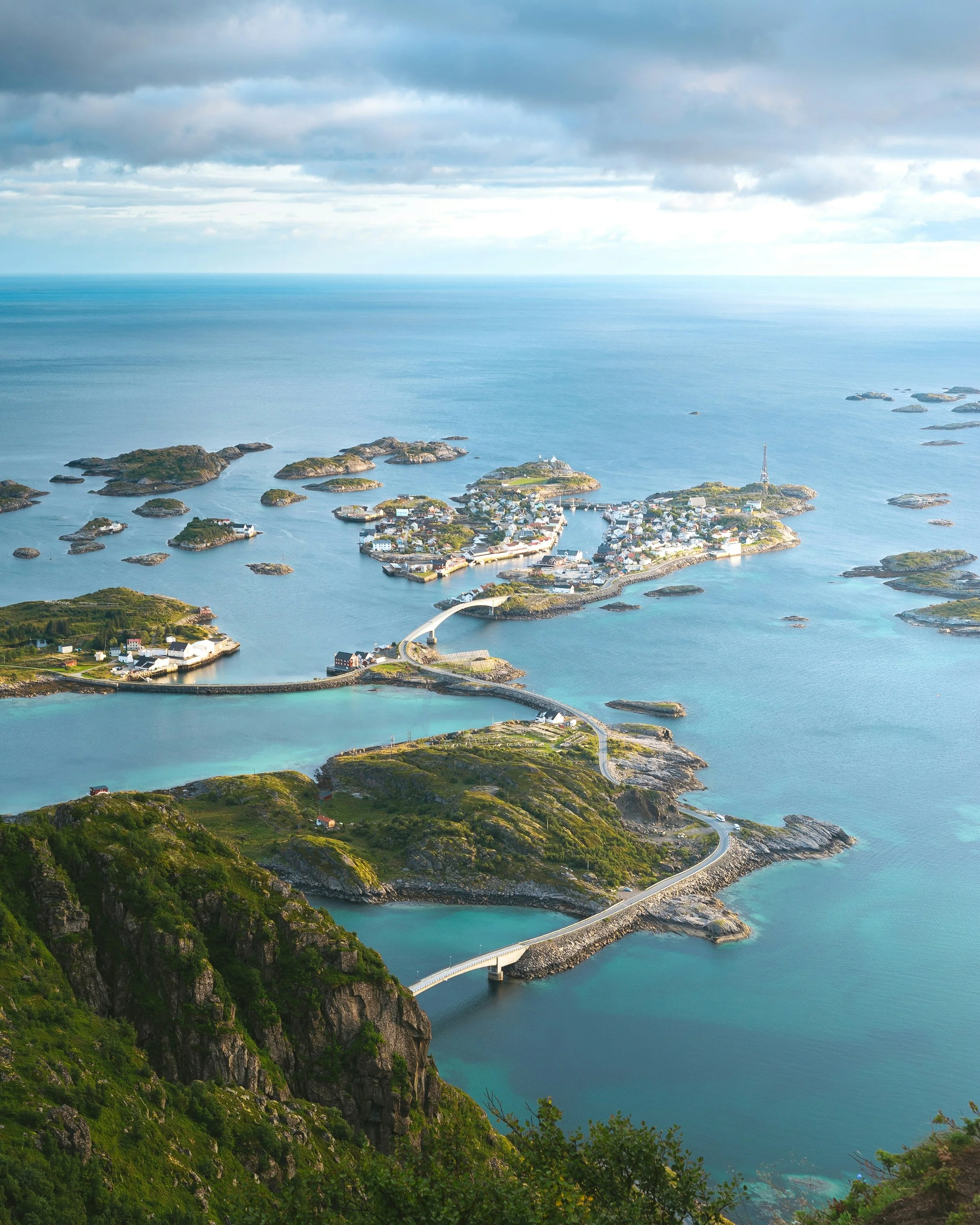 Aerial view of a coastal village with houses, a bridge, rocky islands, green hills, and the sea under a cloudy sky.