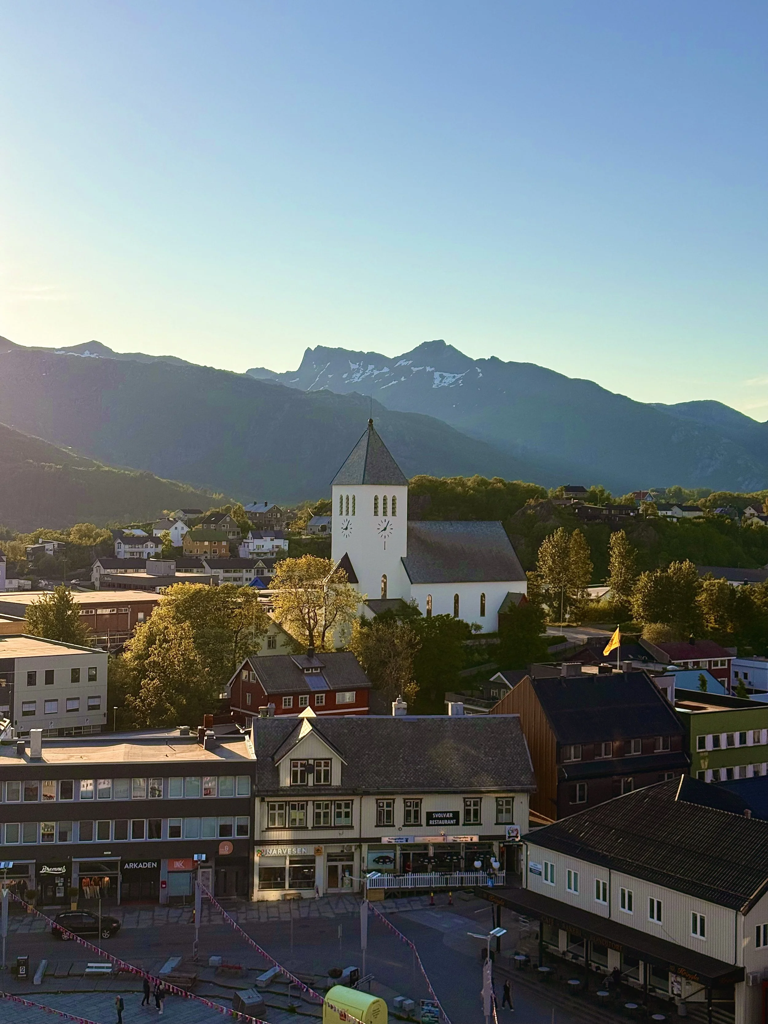 Scenic view of a small town with a white church with a tall steeple, surrounded by houses, with mountains in the background and a clear blue sky.