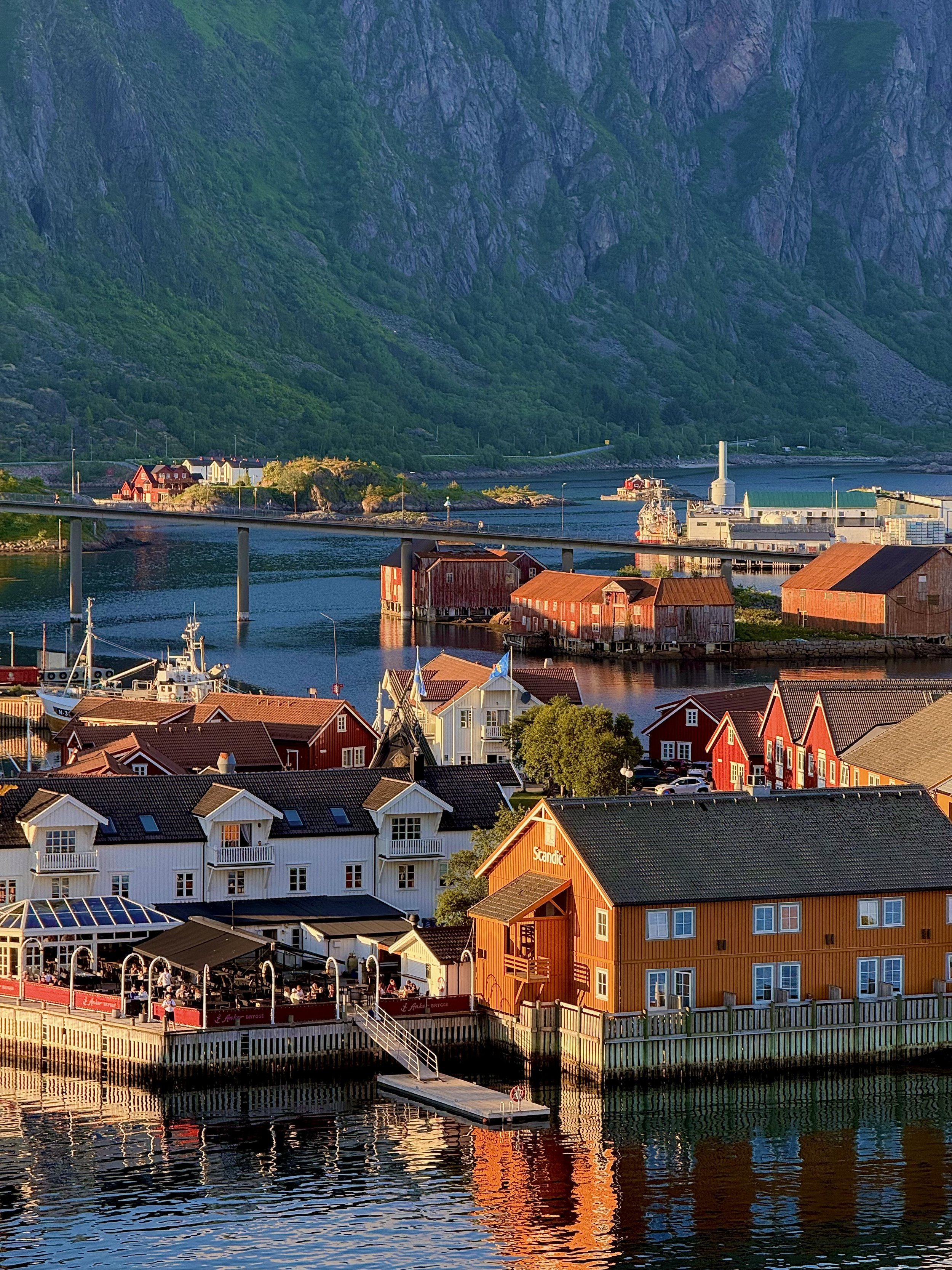 Colorful waterfront village with red, white, and orange buildings, a bridge, boats in the water, green mountains in the background, and a dock reflected in the water.
