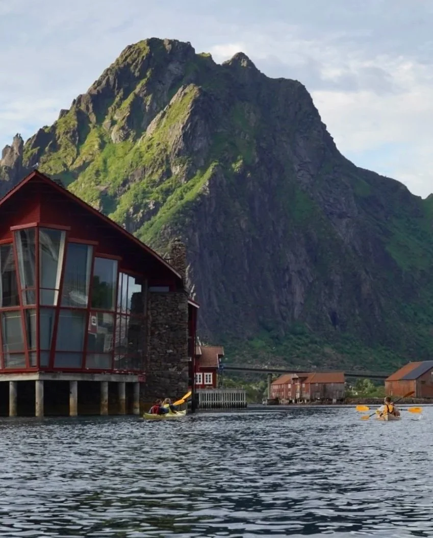 People kayaking in a lake with a modern house on stilts and a mountain in the background on a partly cloudy day.