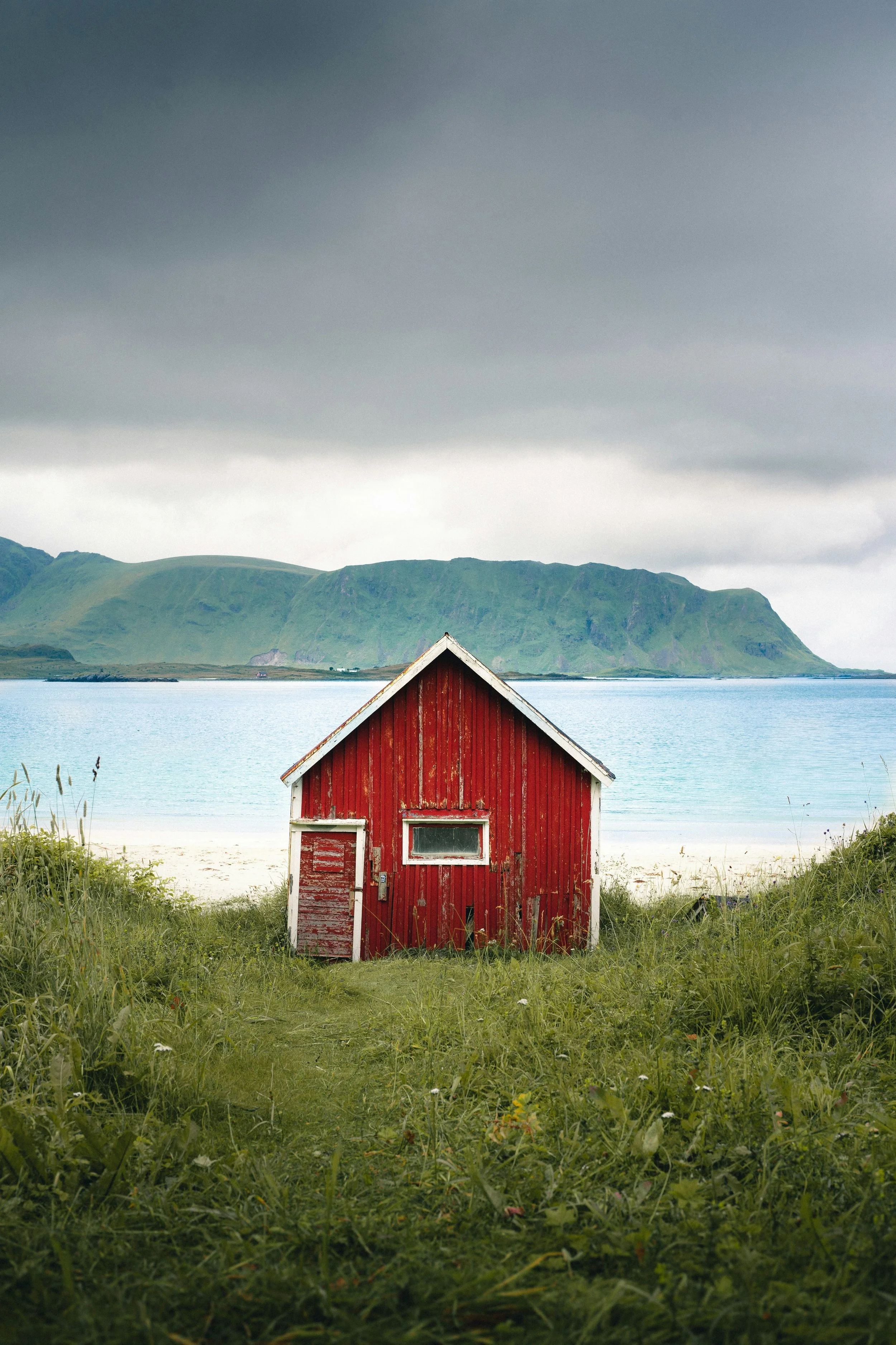 A small red wooden building with peeling paint sits on a grassy area near a body of water. In the background, there are green mountains under a cloudy sky.