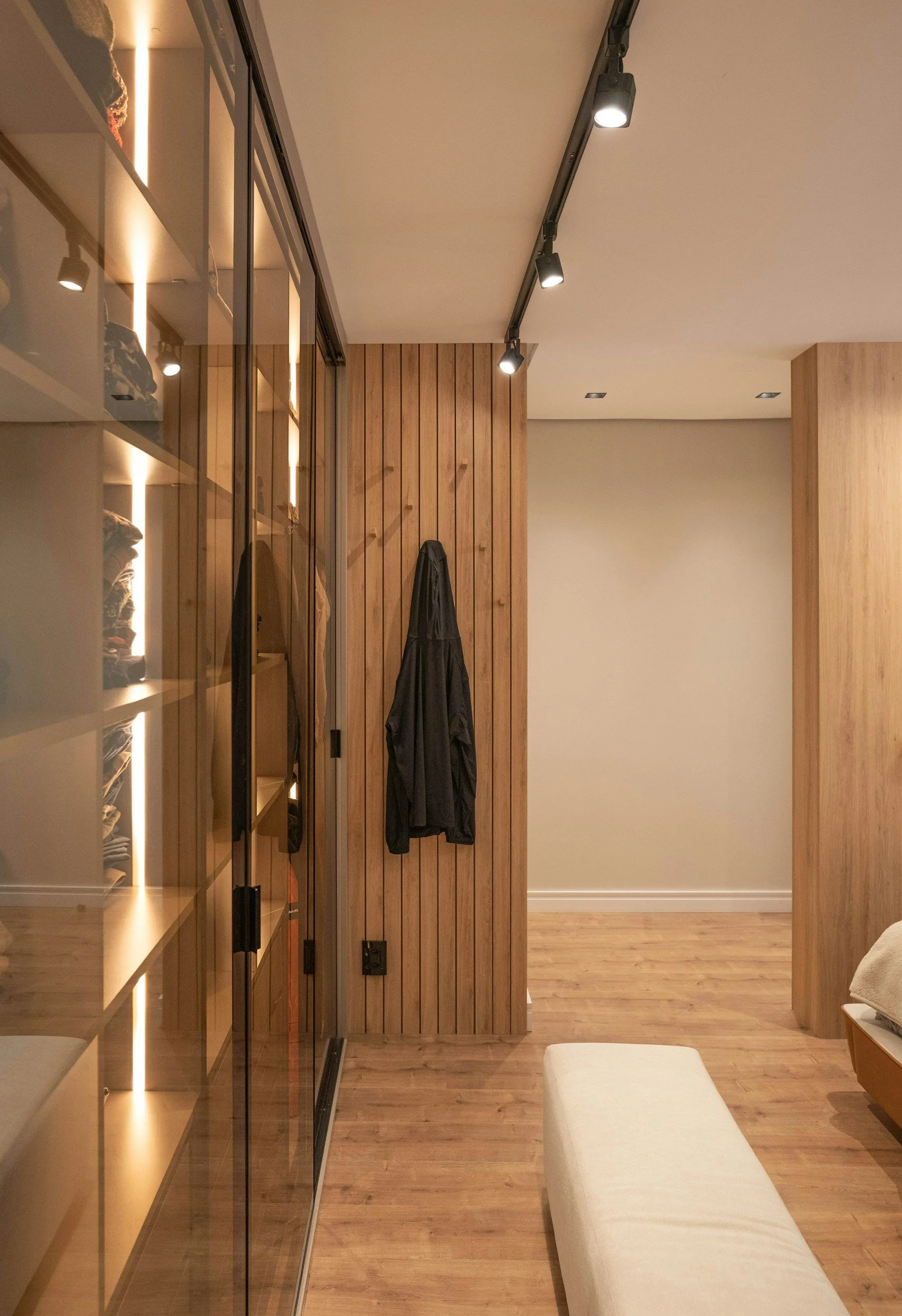 A modern interior space with wooden wall paneling, track lighting on the ceiling, a glass-fronted closet or storage area, a black coat hanging on the wood wall, and a beige upholstered bench on a wooden floor.