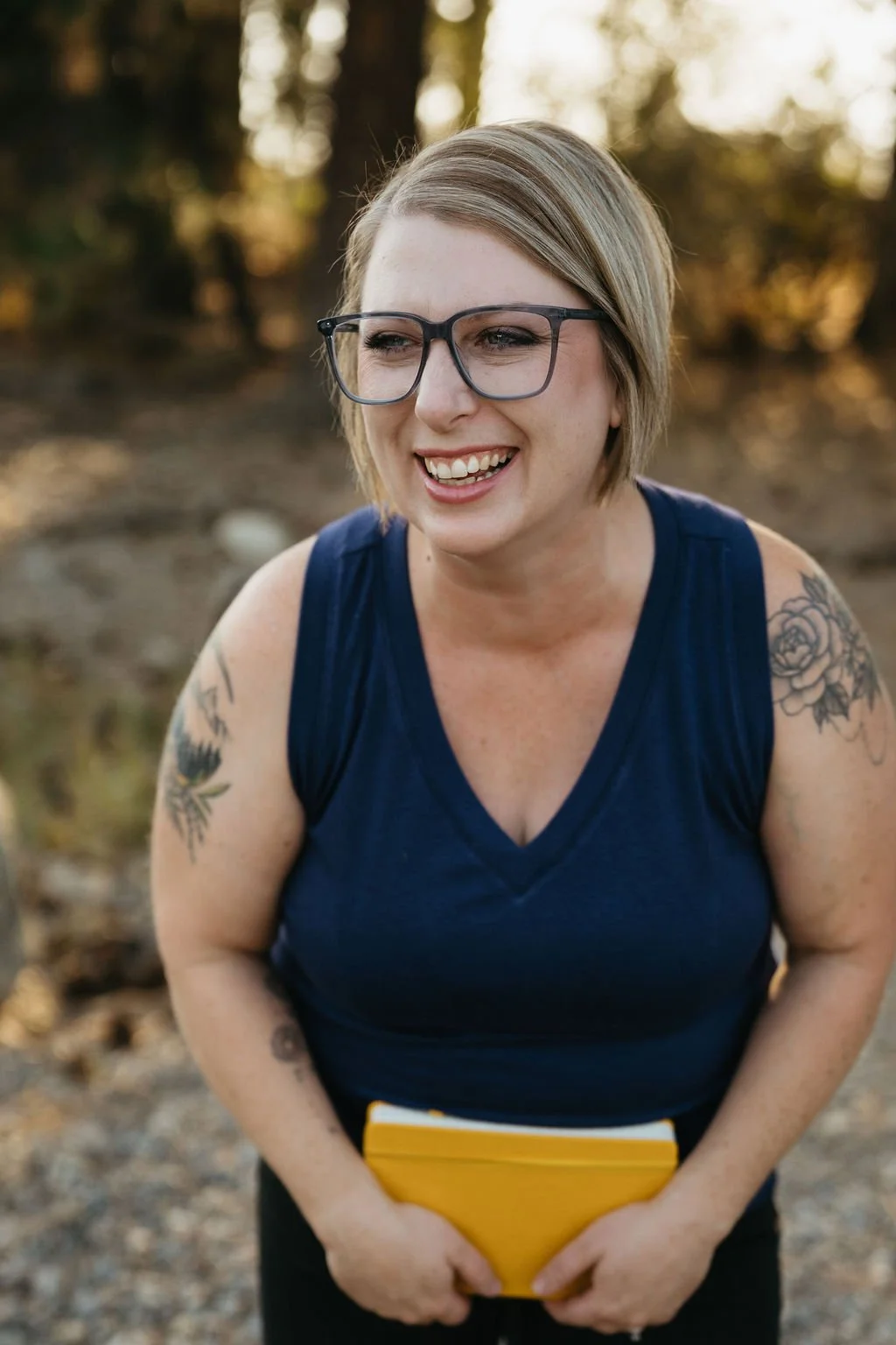 A woman with short blonde hair, glasses, and tattoos on her arms, wearing a sleeveless navy blue top, outdoors with trees in the background, smiling and holding a yellow notebook.