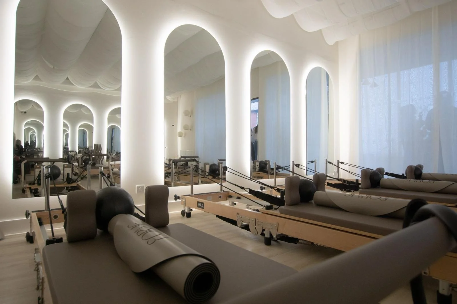 Pilates or fitness studio with large mirrors, arched windows with sheer curtains, and multiple reformer Pilates machines.