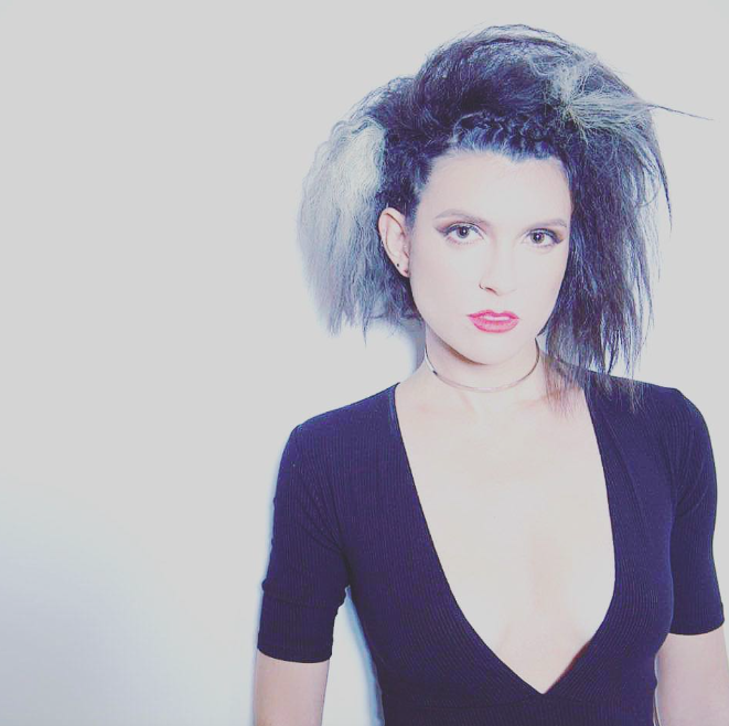 A woman with messy, voluminous black and gray hair, wearing a deep V-neck black top, and a choker necklace, standing against a plain light background.