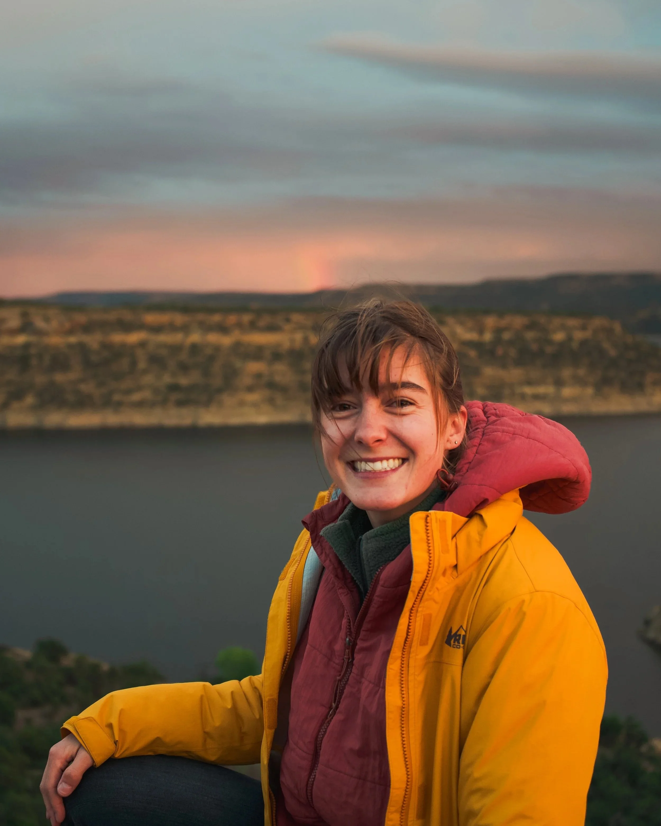 Portrait of a smiling young woman with short brown hair wearing a yellow jacket and a maroon hoodie, outdoors during sunset with a blurred landscape in the background.