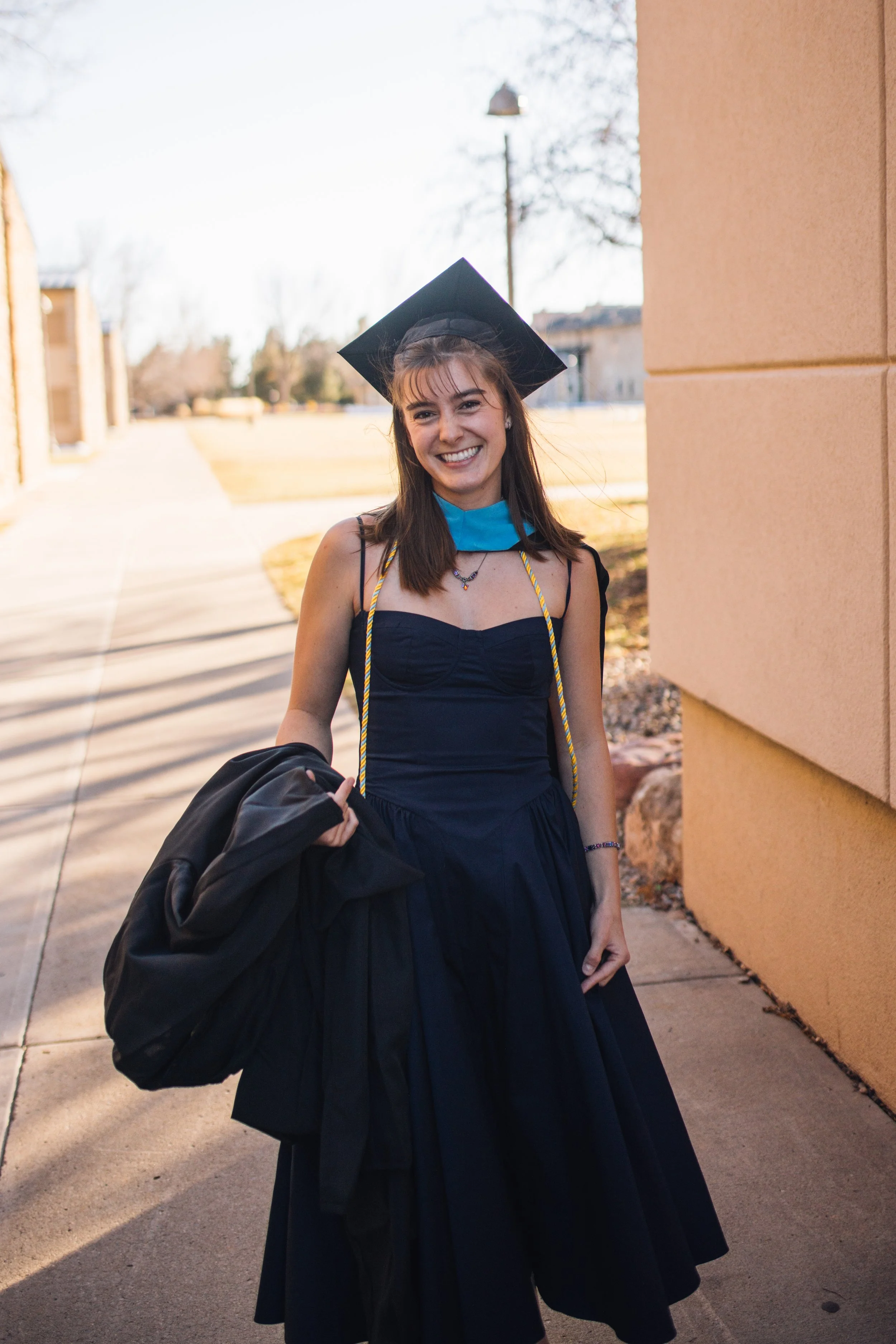 A young woman in a graduation cap and gown standing outdoors, smiling and holding a jacket, with a building and trees in the background.