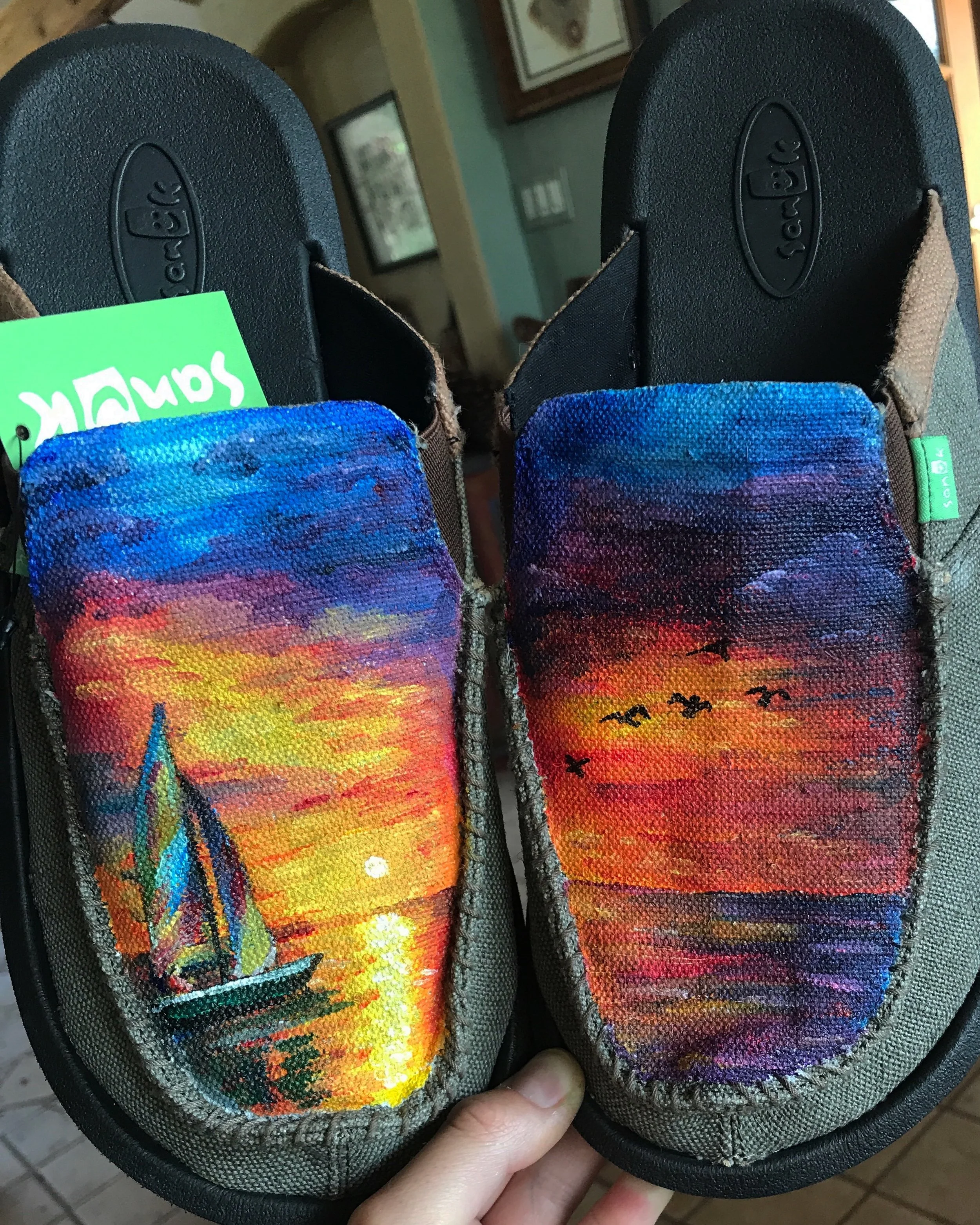 The image shows the top of a pair of shoes with a colorful sunset and sailboat hand painted on the fabric part near the toe area.