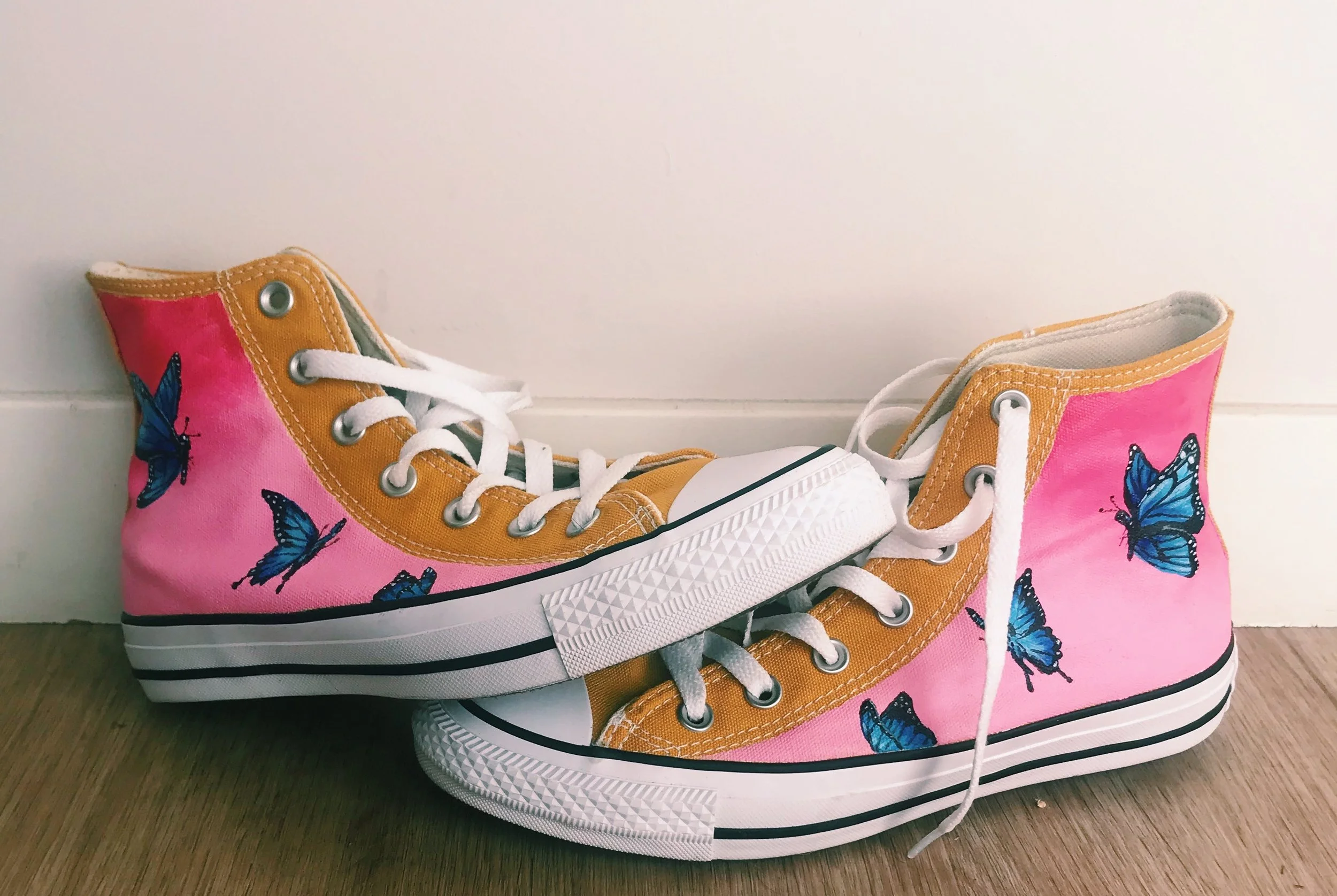 Pair of pink high-top canvas sneakers hand painted with blue butterflies. the butterflies are painted realistically on the side of the shoe. 