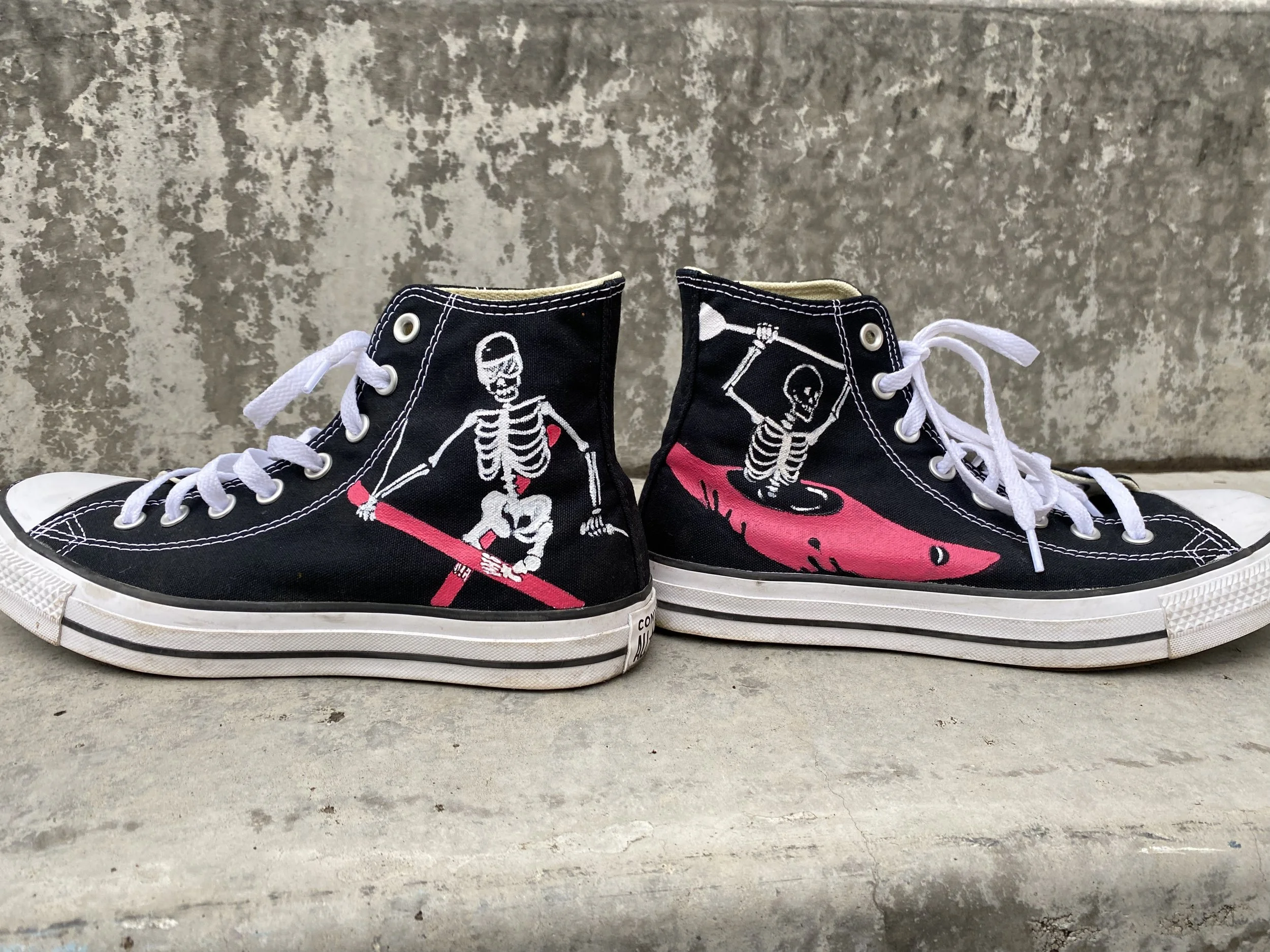 A pair of black high-top canvas sneakers with hand painted skeletons. The skeletons are in a white water kayak and doing a trick on skis.