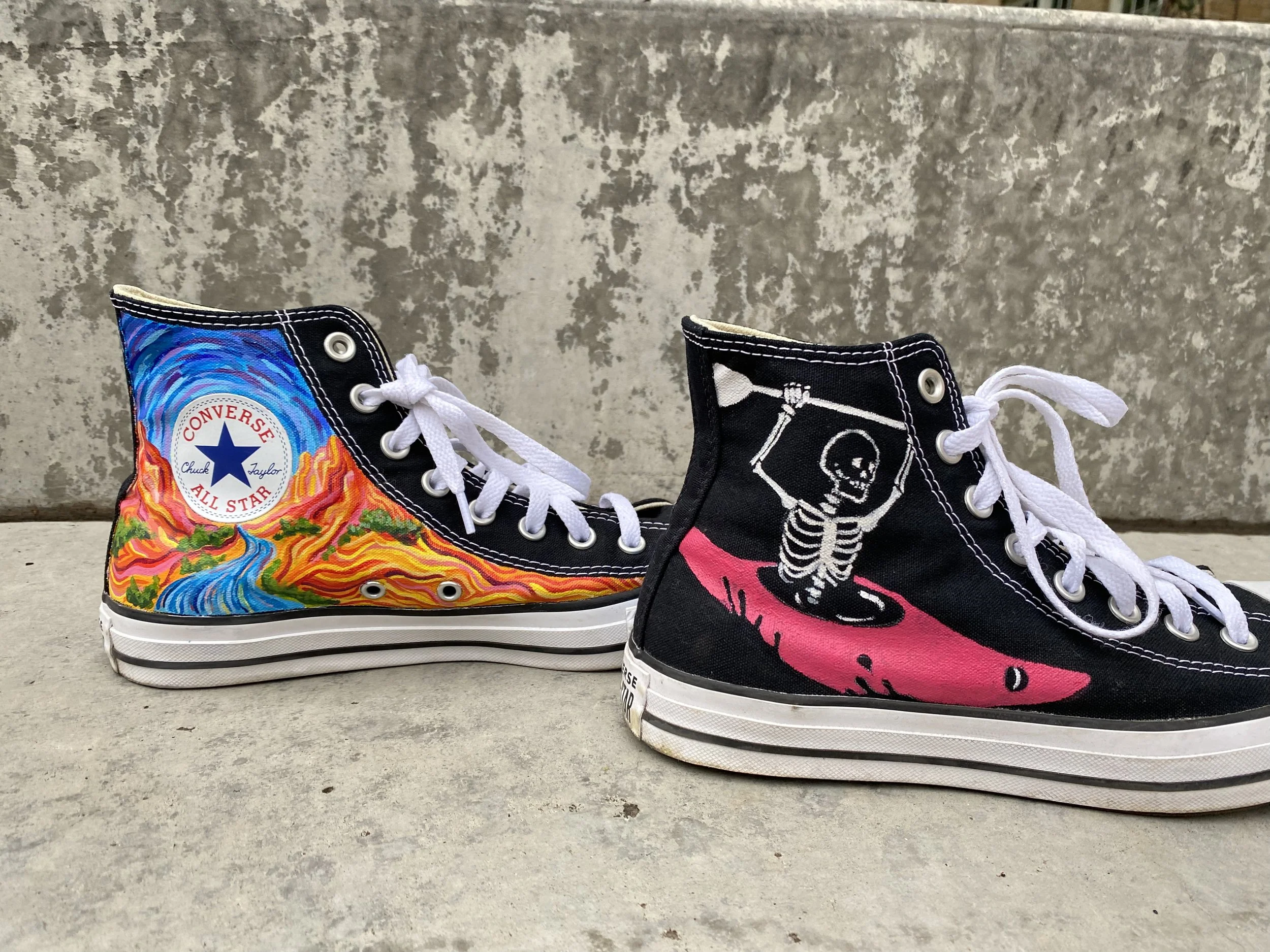 Two high-top Converse sneakers with hand painted designs, featuring a skeleton in a whitewater kayak, and a desert river scene. 