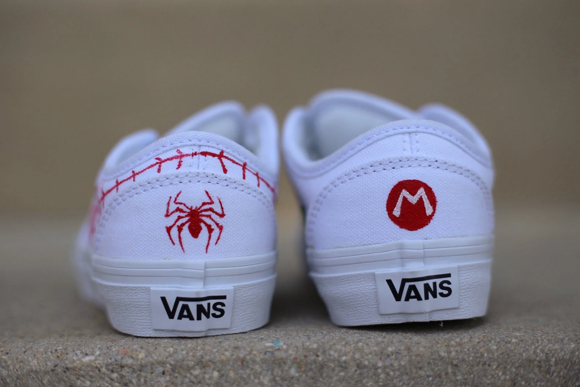 Back of white Vans sneakers hand painted with red Spider-Man themed designs. The left shoe features a red spider web and a spider, while the right shoe has a red Super Mario Bros symbol.  
