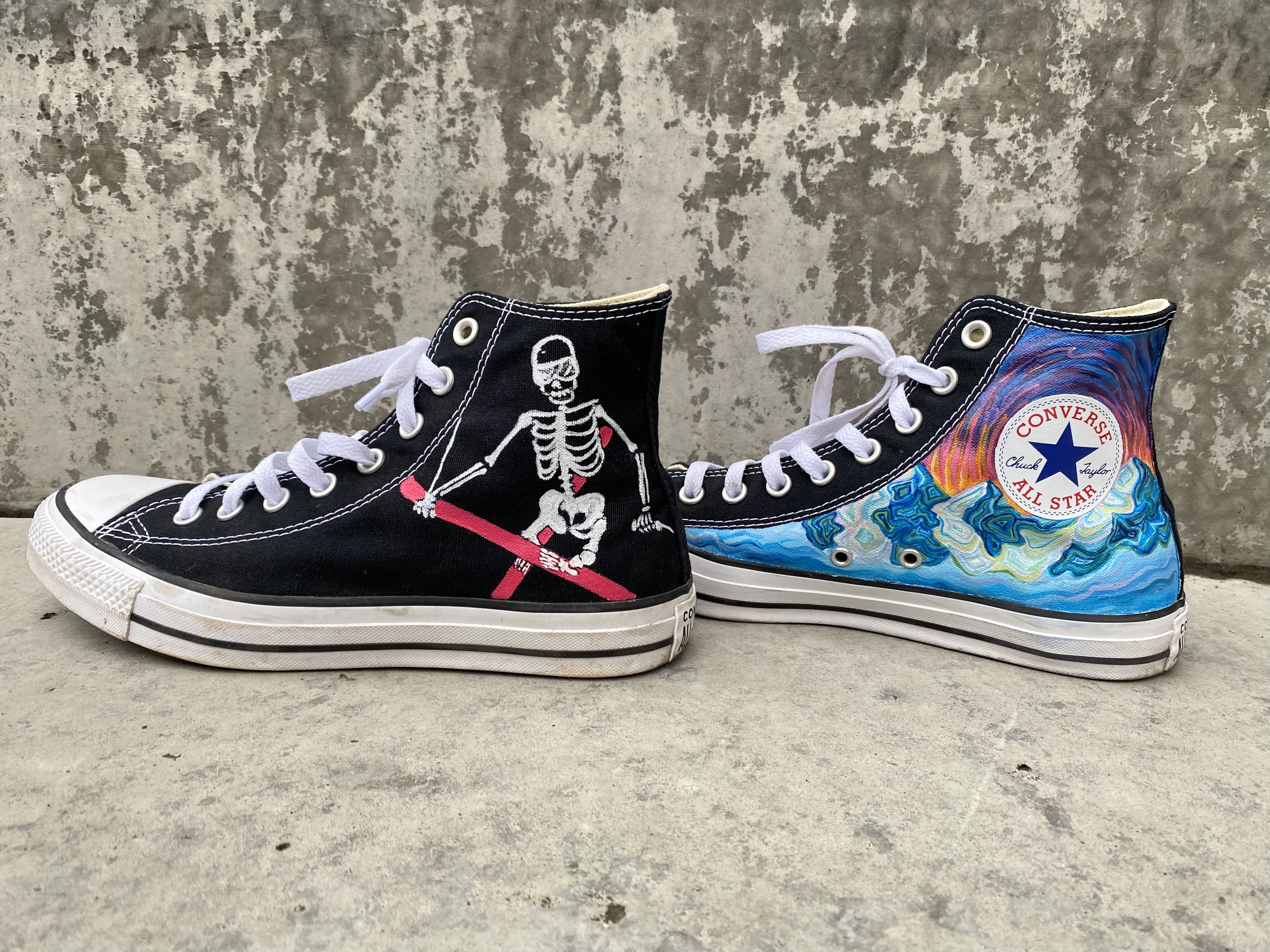 Pair of black high-top Converse sneakers with hand painted artistic designs, including a skeleton wearing skis on the left shoe and blue and multicolored snowy mountain scene on the right shoe. 