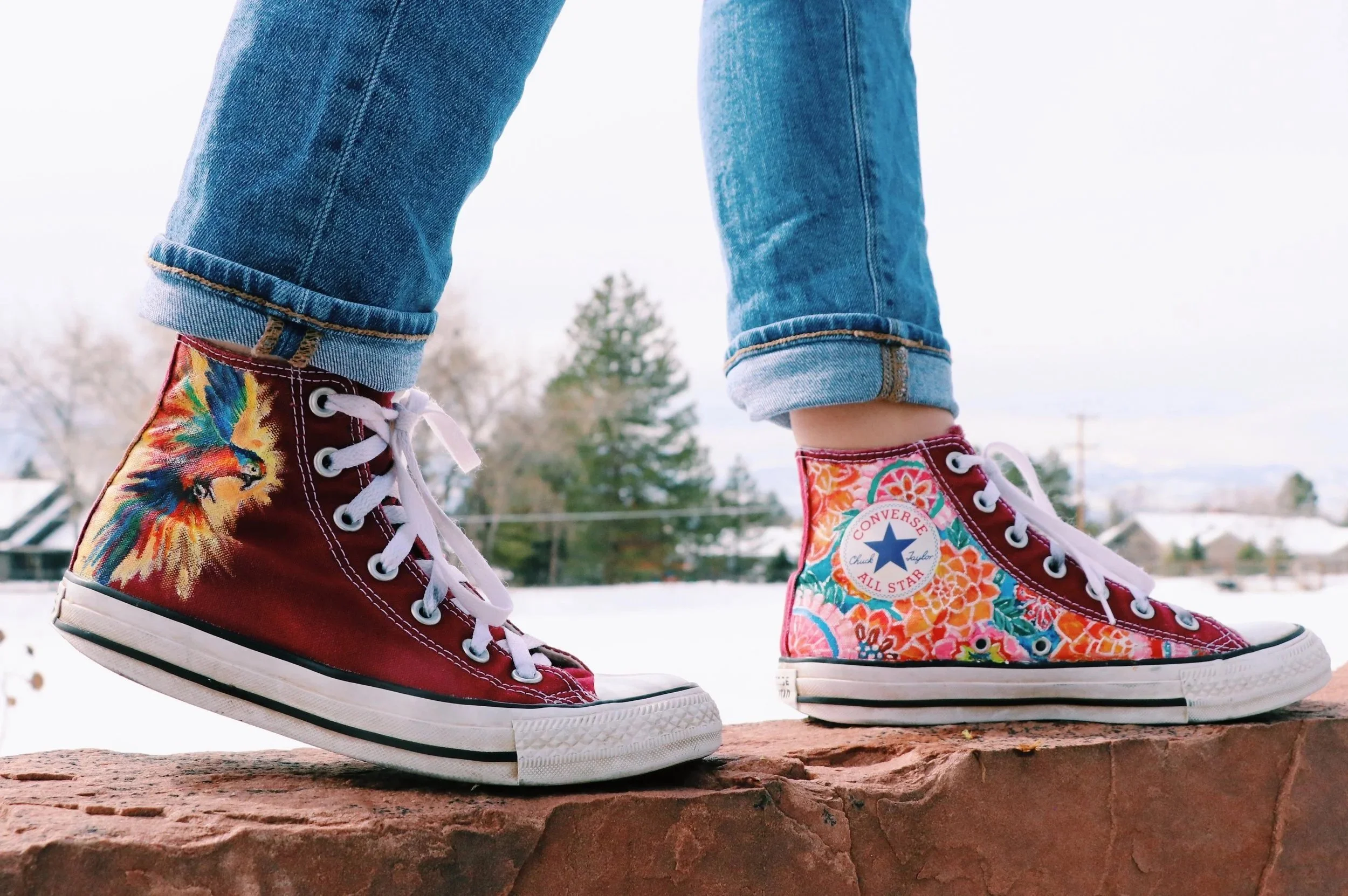 Close-up of colorful high-top Converse sneakers, painted with a rainbow floral design, and a parrot on the side of the shoe. The shoes are handpainted.