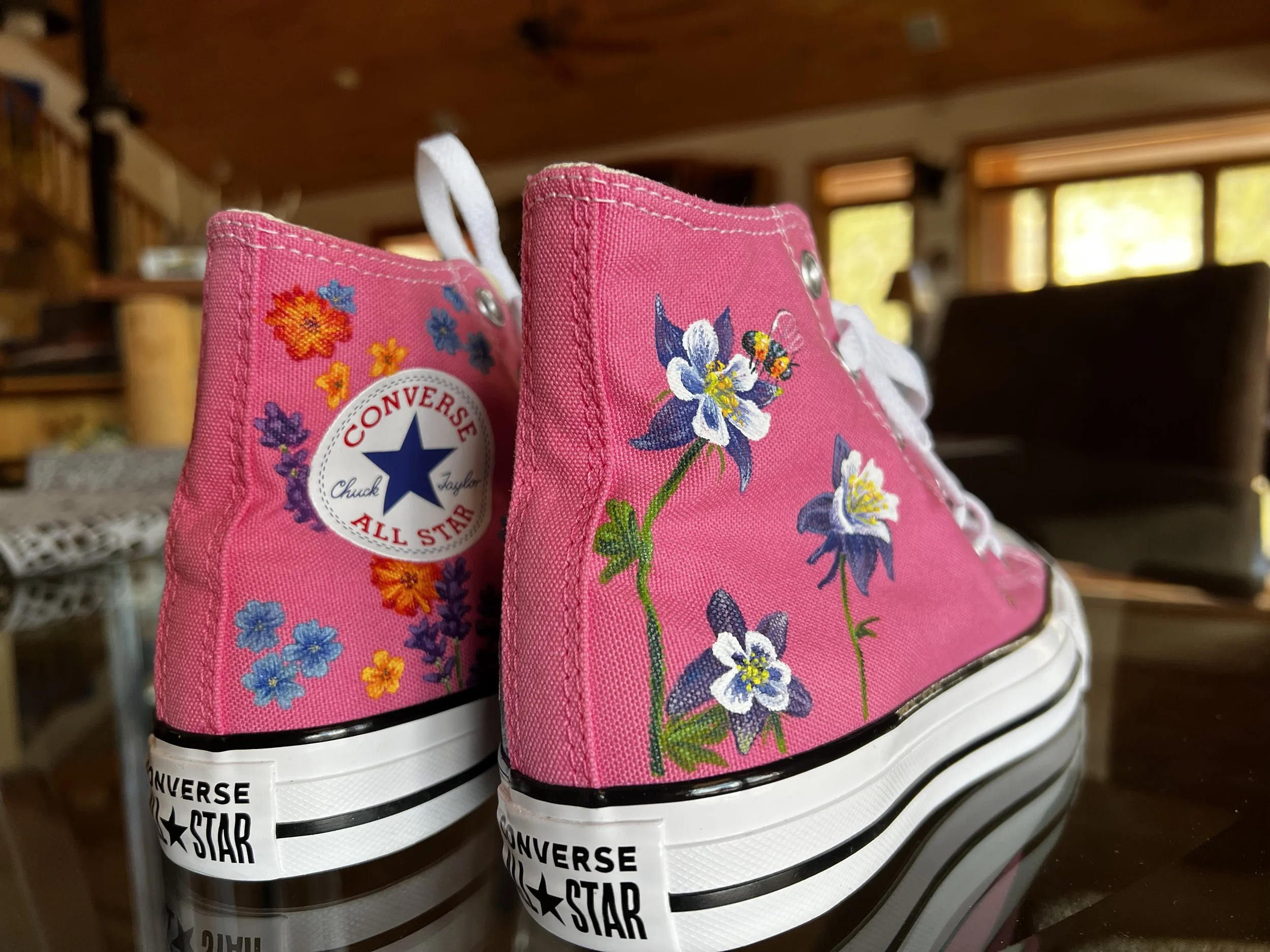 Pink Converse Chuck Taylor All Star sneakers with floral hand painted designs, including columbine flowers and a bee on one shoe. The other shoe is hand painted with colorful wildflowers.