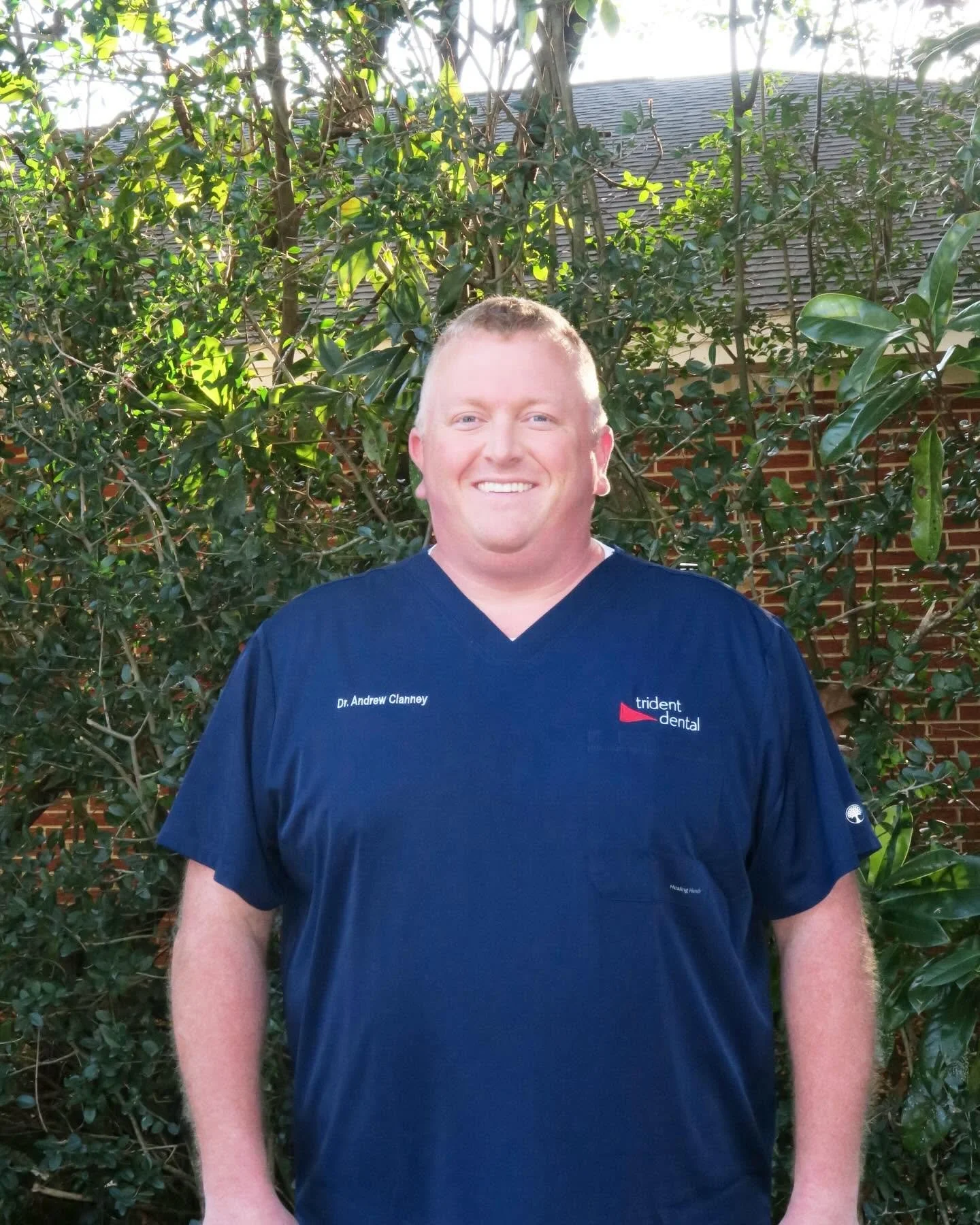 A smiling man with short blond hair wearing a navy blue medical scrubs top standing outdoors in front of green bushes and trees with a house and brick wall in the background. Dental practice sales testimonial and review