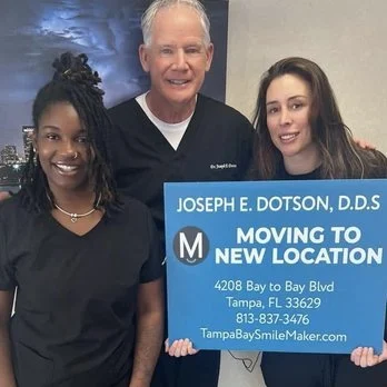 Three individuals standing together holding a sign that reads 'Joseph E. Dotson, D.D.S. Moving to New Location, 4208 Bay to Bay Blvd, Tampa, FL 33629, 813-837-3476, TampaBaySmileMaker.com' in front of a plain background. Dental practice sales testimo
