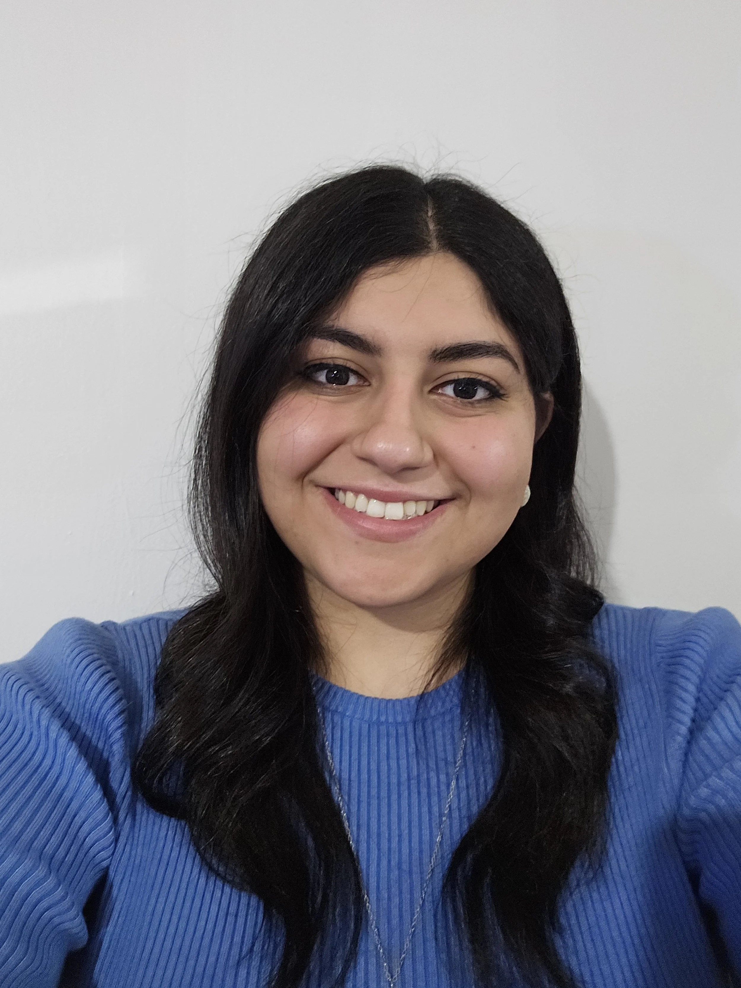 A woman with long dark hair smiling in front of a plain white wall, wearing a blue ribbed sweater.