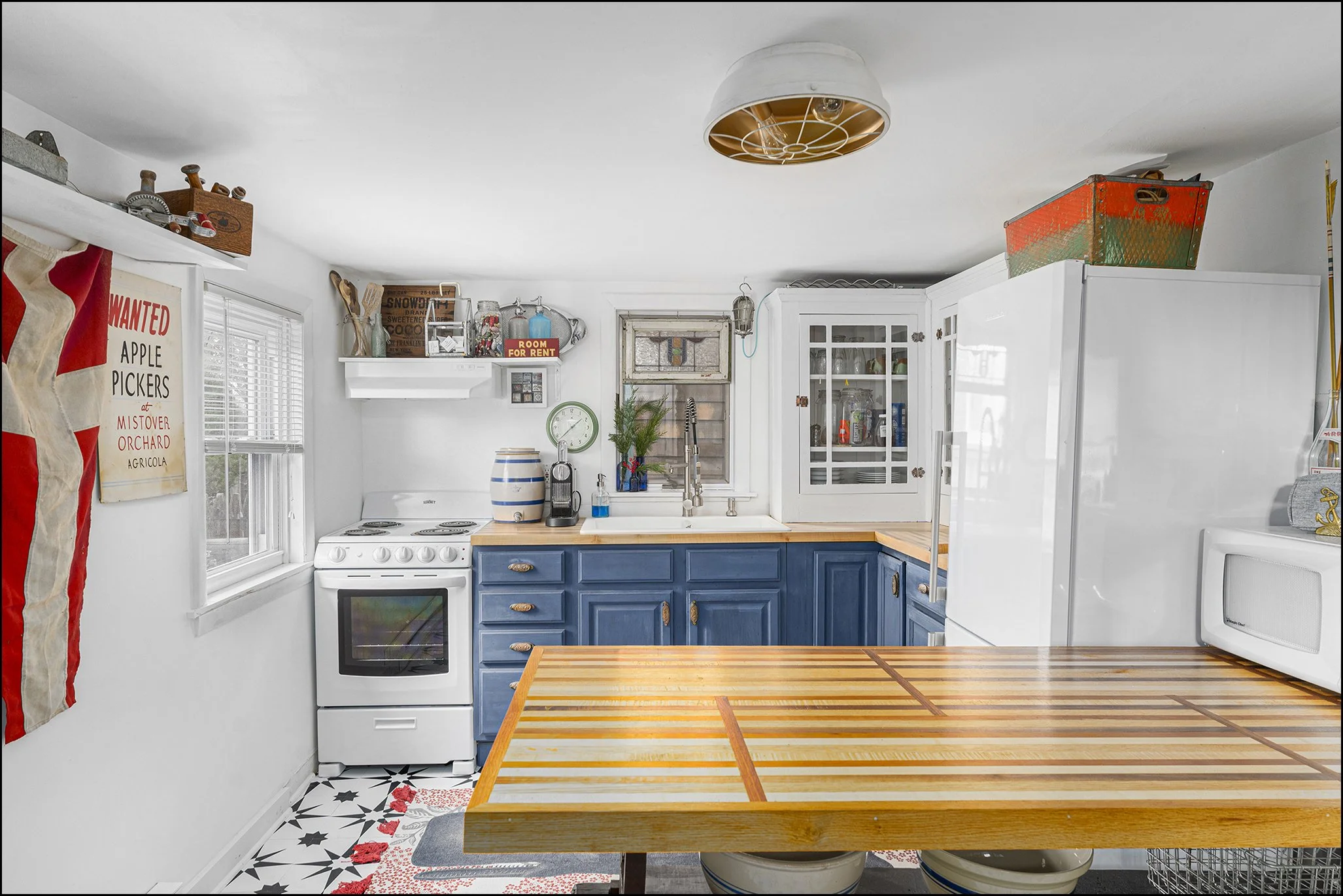 A kitchen with white walls and cabinets, blue lower cabinets, a wooden table, a white stove, a microwave, a window, shelves with various items, and a colorful storage basket on top of the cabinets.
