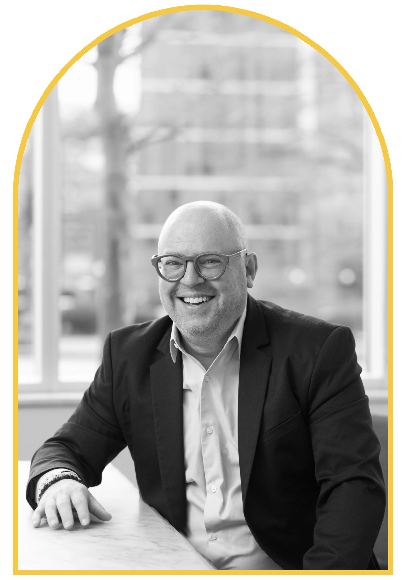 Portrait of a bald man wearing glasses and a suit, smiling, sitting at a table with a window in the background.