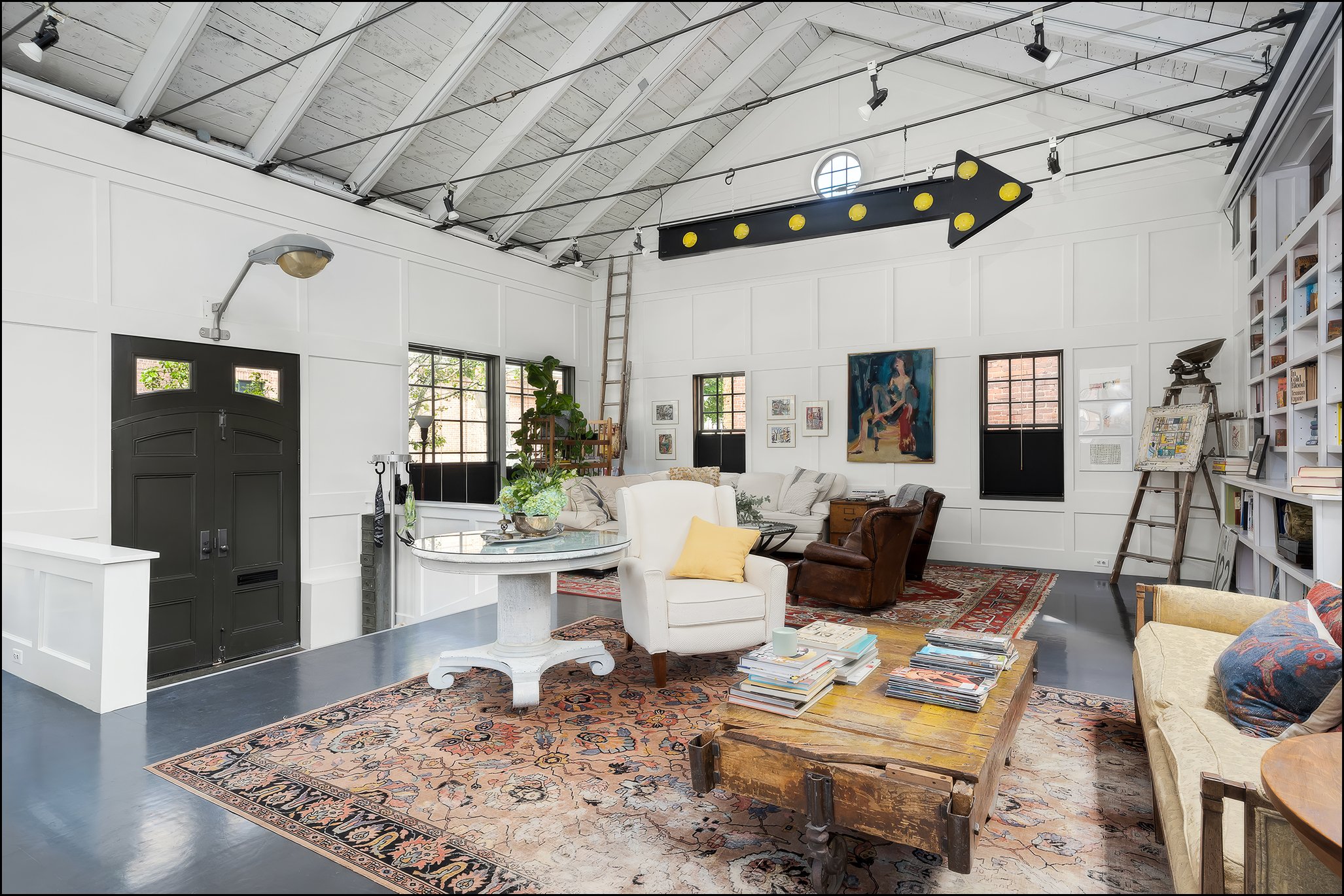 A spacious living room with white paneled walls, large windows, and a wooden vaulted ceiling. The room is decorated with various artworks, books, and furniture including a white armchair with a yellow pillow, a leather armchair, and a wooden table. T