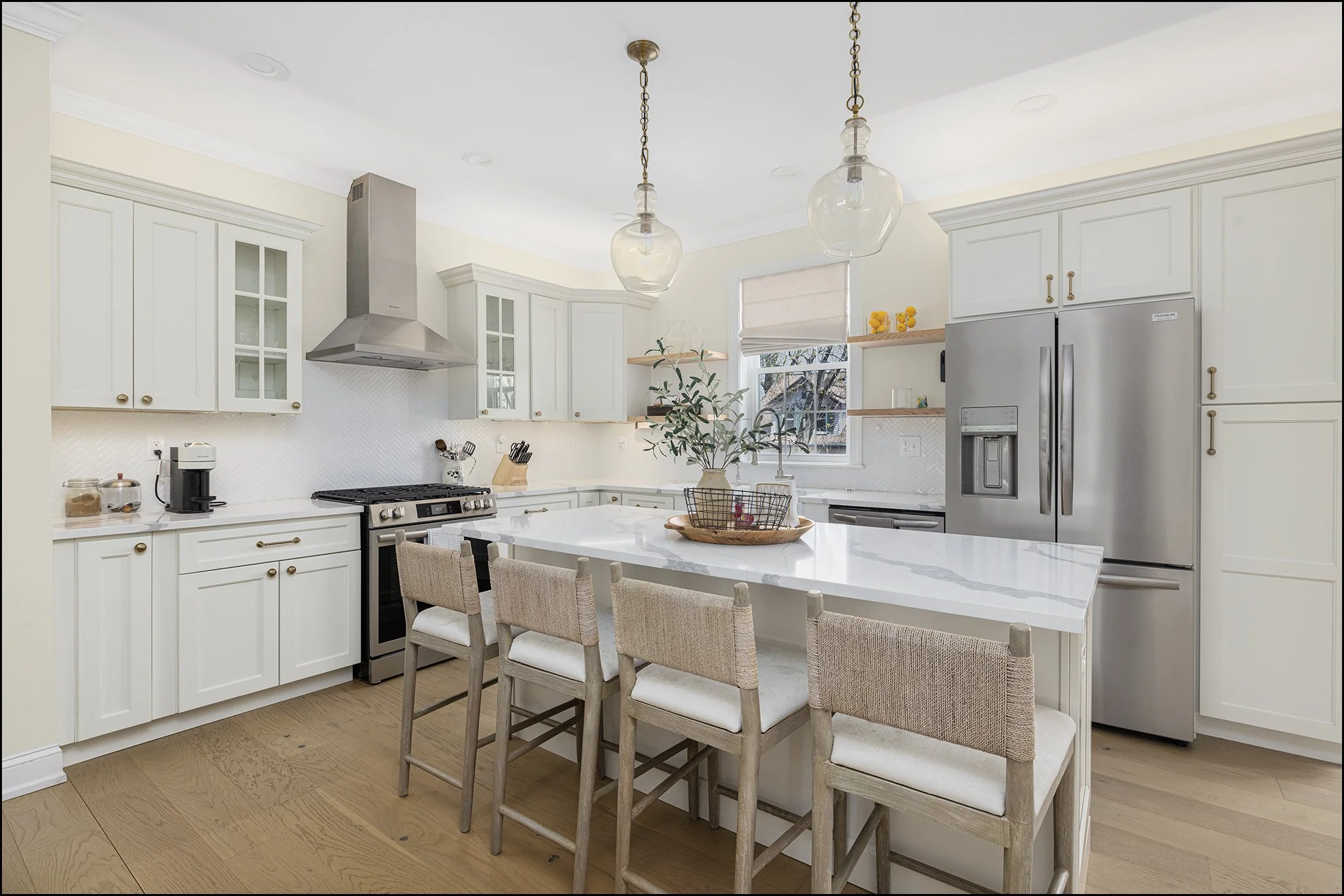 Bright kitchen with white cabinetry, stainless steel refrigerator, gas stove, and kitchen island with chairs. Hanging pendant lights, wooden open shelves, and a window with blinds above the sink.