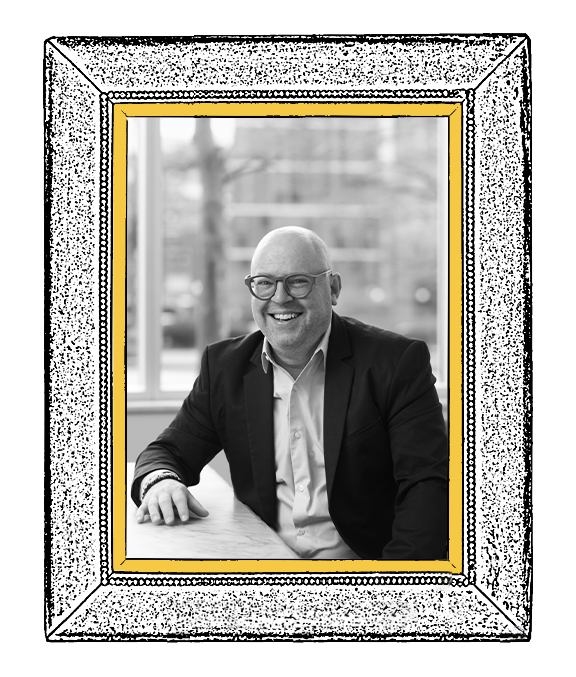 A black and white photo of a smiling, bald man wearing glasses, a white shirt, and a dark blazer, sitting at a desk in front of a window with a blurred outdoor view.