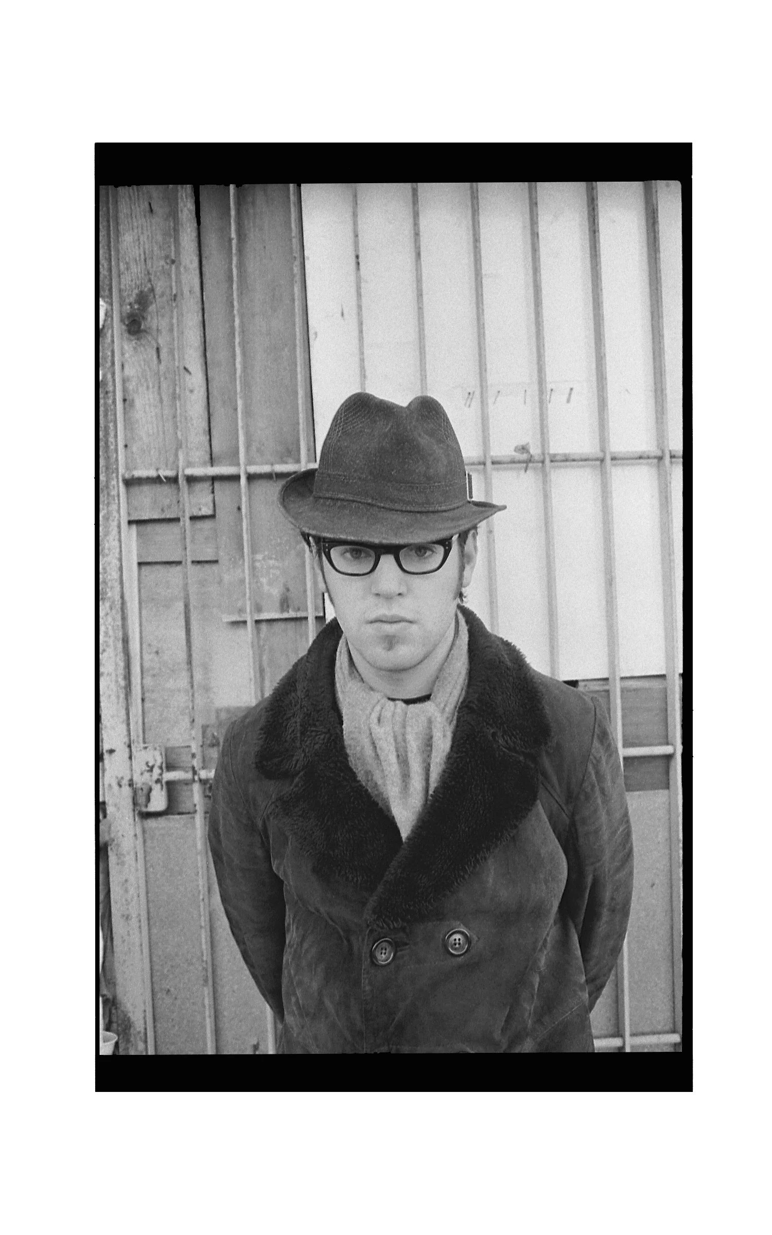 A black-and-white photo of a young man wearing glasses, a fedora hat, a coat with a fur collar, standing in front of a wooden fence or gate.