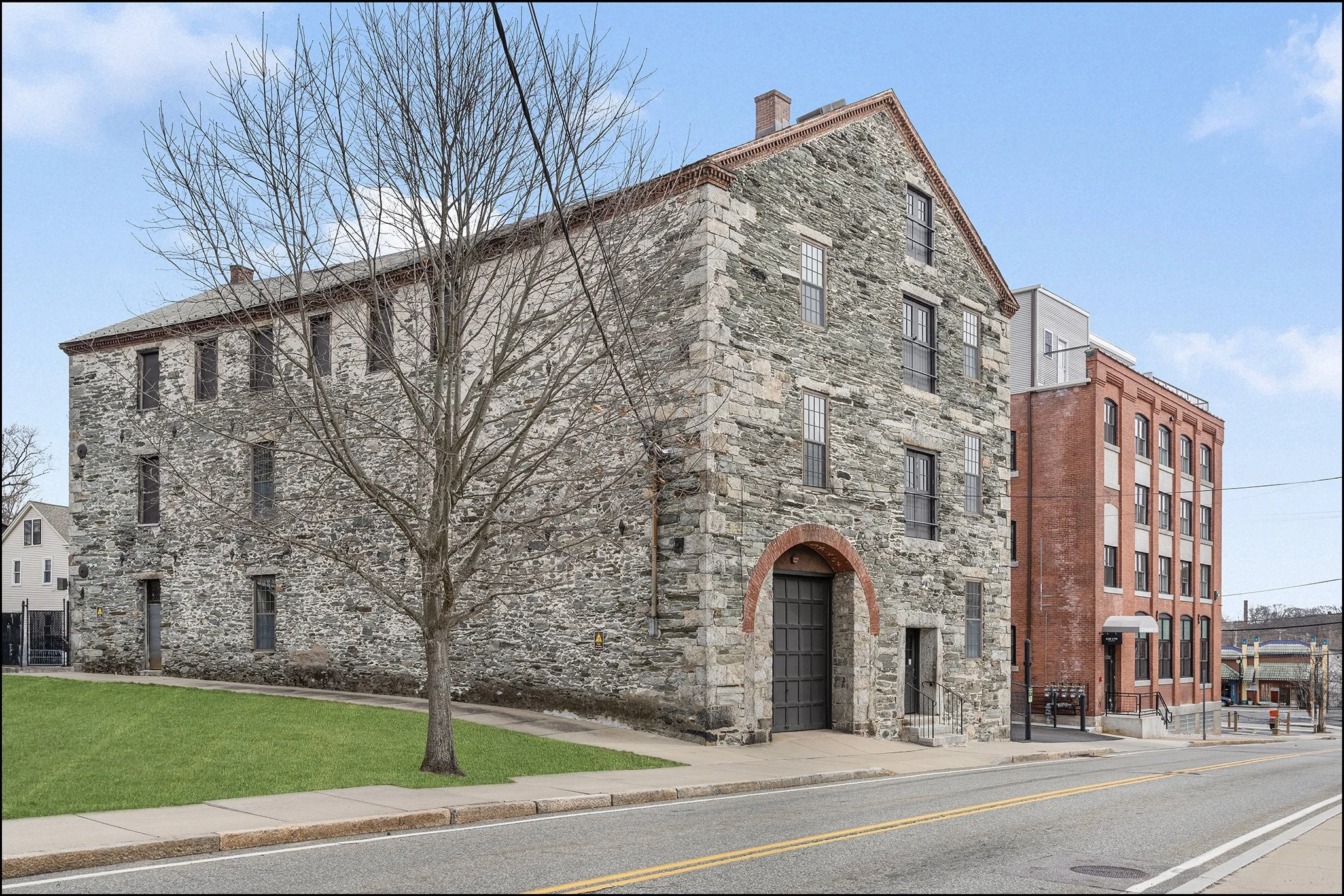 A street view featuring a large old stone building with arched windows and a black door, alongside modern brick and metal buildings. There is a leafless tree and a green grassy area in front of the stone building, with a sidewalk and street in the fo