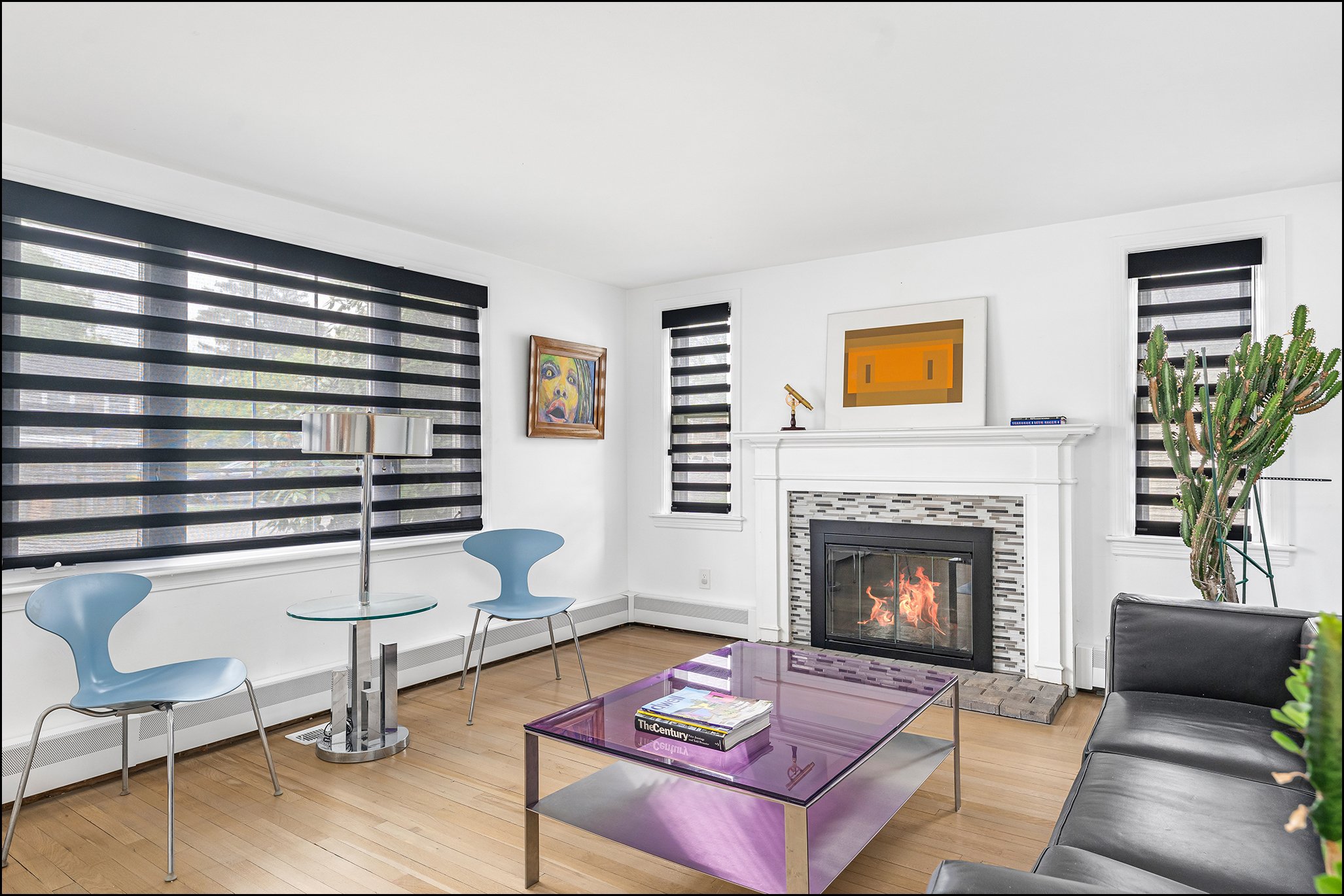 A modern living room with black striped window blinds, wall art, a fireplace, a cactus plant, and a purple glass coffee table.