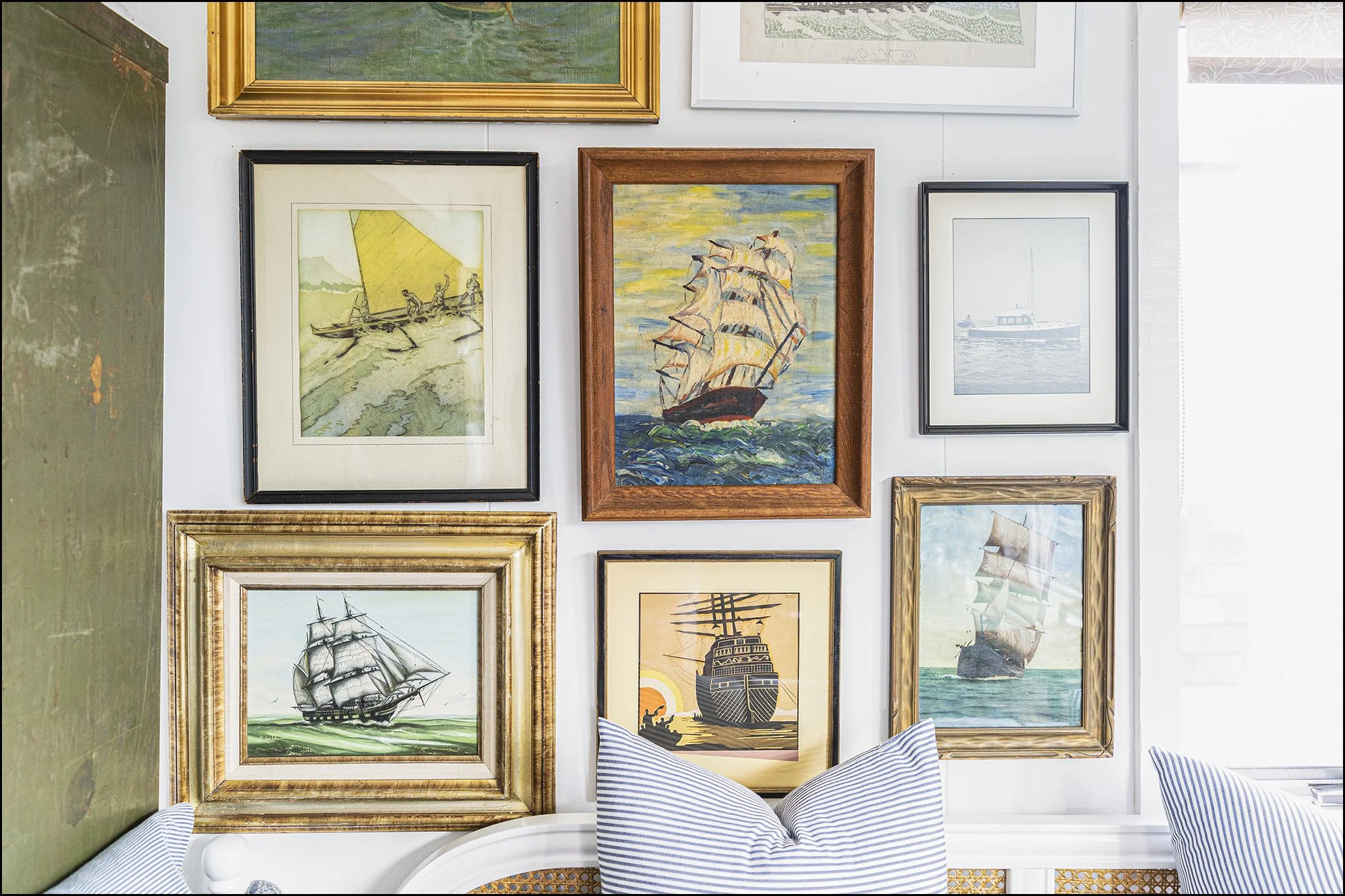 A wall with multiple framed paintings of ships, boats, and sea scenes, with parts of a striped pillow visible at the bottom.