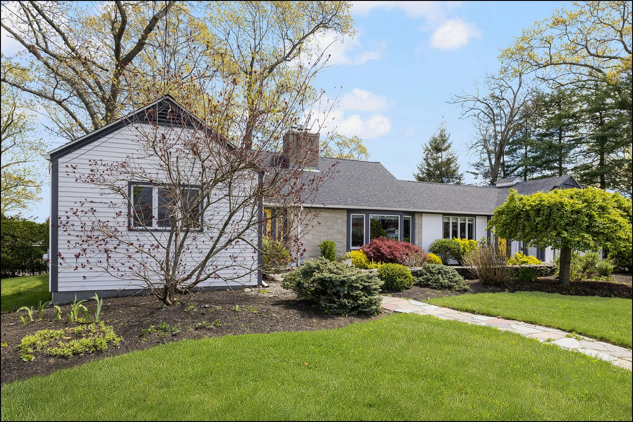 Front yard of a house with a well-maintained lawn, various shrubs and trees, and a sidewalk leading to the house. The house has white siding, large windows, and a gray shingled roof.
