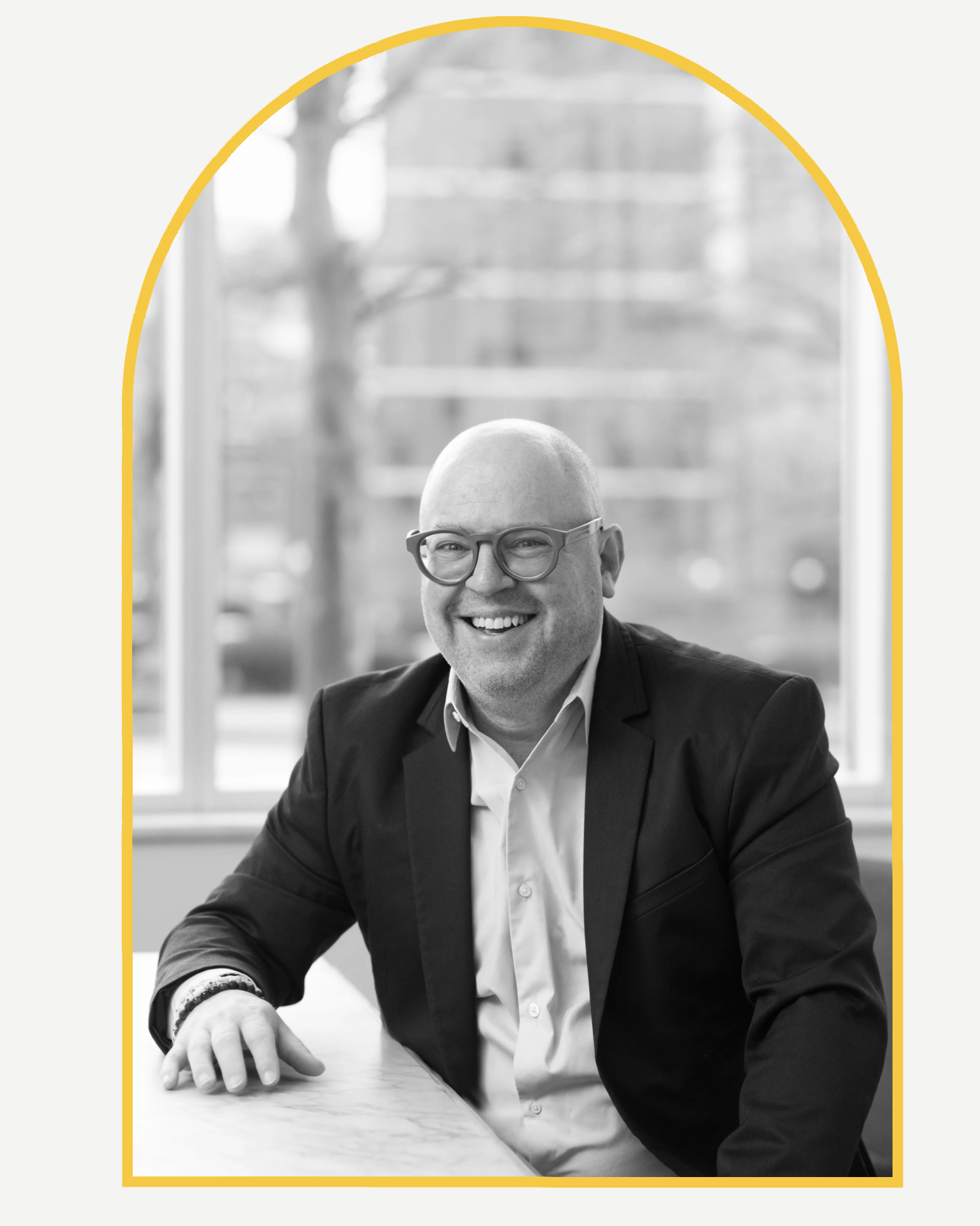 A smiling man with a bald head and glasses wearing a suit and white shirt, sitting at a table with a blurred background outside a window.