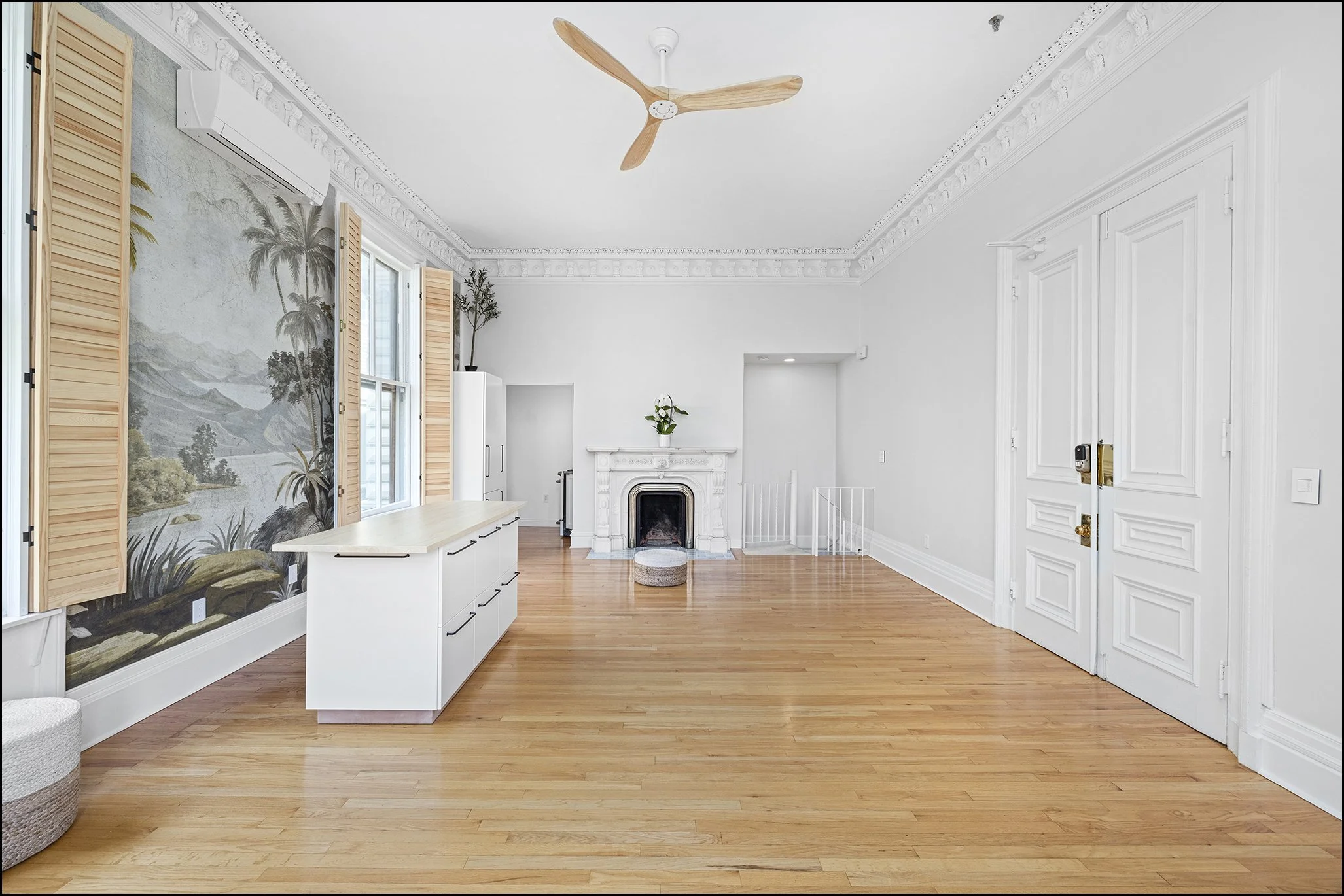 Living room with hardwood floors, white walls, decorative crown molding, and a ceiling fan. There is a fireplace with a white mantel, a small potted plant on top, and a large window with light wood shutters. The room has minimal furniture and decor, 