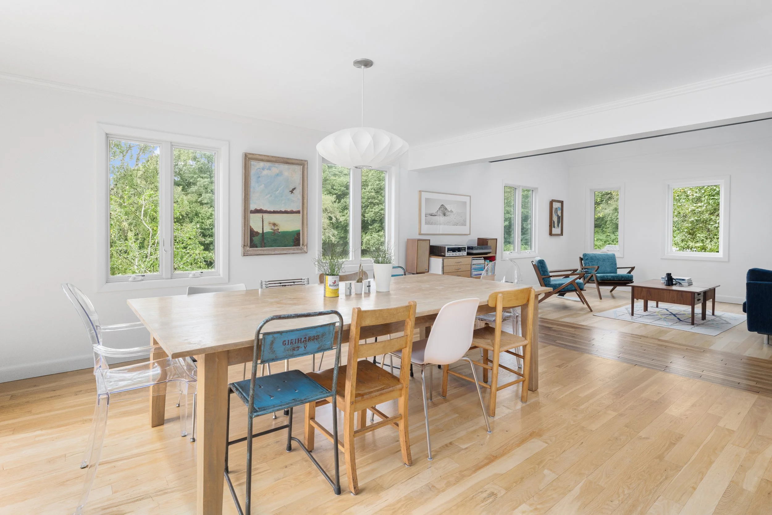 A bright, airy living room with a large wooden dining table, mismatched chairs, and a cozy seating area with blue armchairs. Large windows show green trees outside, and framed artwork decorates the white walls.