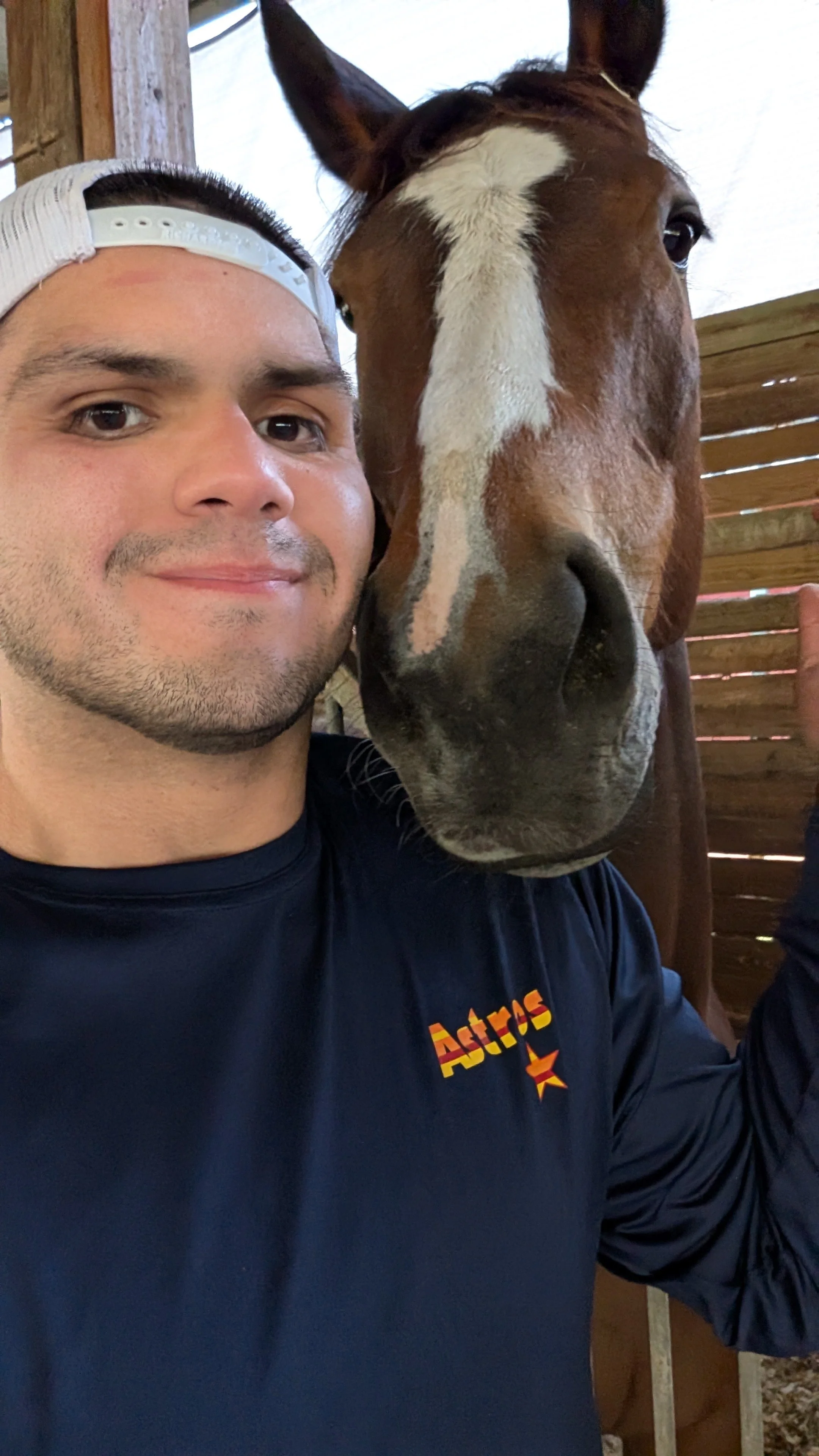 A man with a light beard and wearing a white cap backwards takes a selfie with a brown and white horse in a wooden stable.