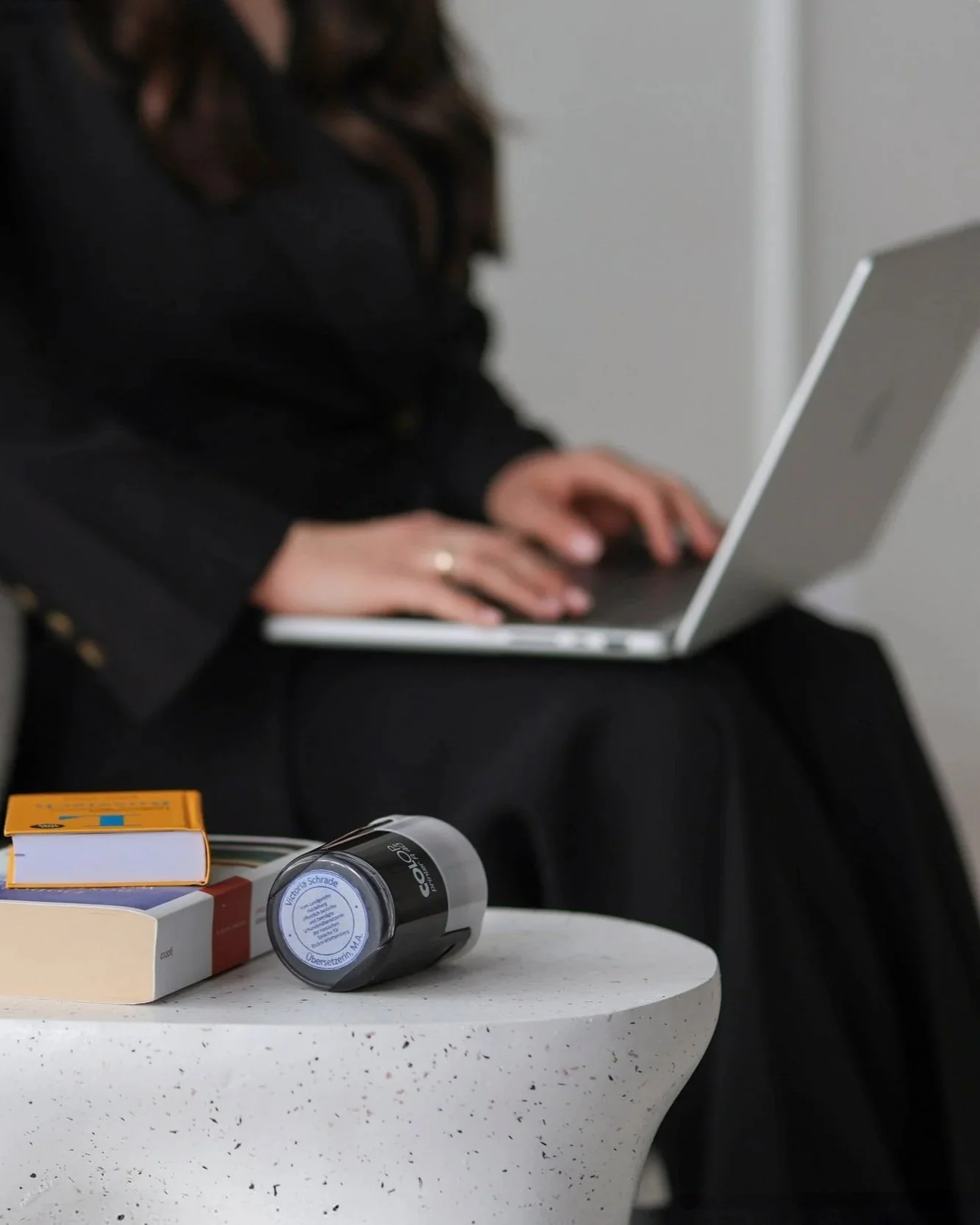 Eine Person sitzt an einem Tisch und arbeitet am Laptop, auf dem Tisch liegen Bücher, eine Schachtel und eine Glühbirne. Im Hintergrund sind weitere Gegenstände sichtbar.