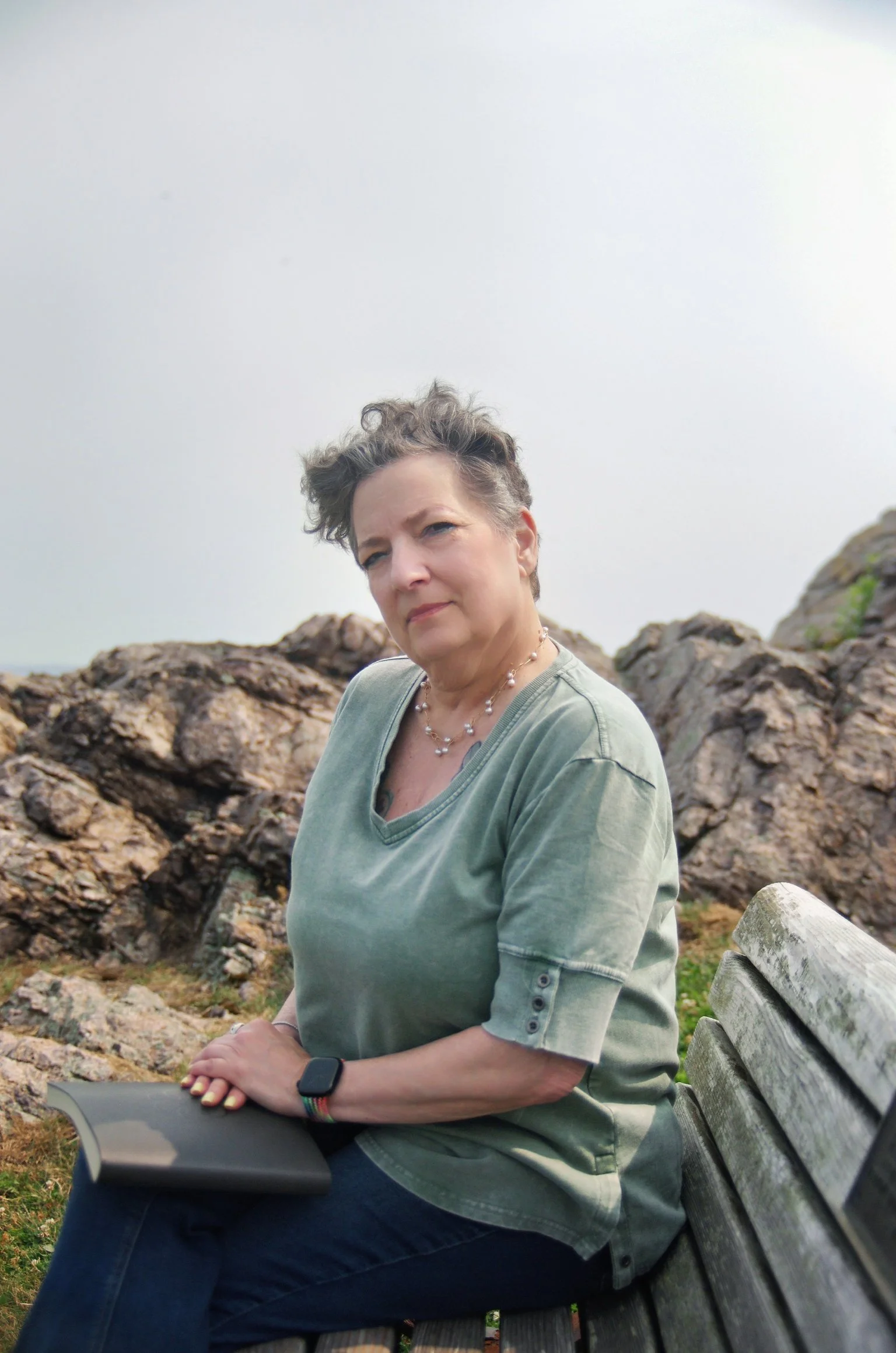 A woman with short, curly gray hair sitting on a park bench outdoors. She is wearing a light green shirt, a necklace, and a smartwatch. She appears to be resting or enjoying the scenery, with rocks and a cloudy sky in the background.