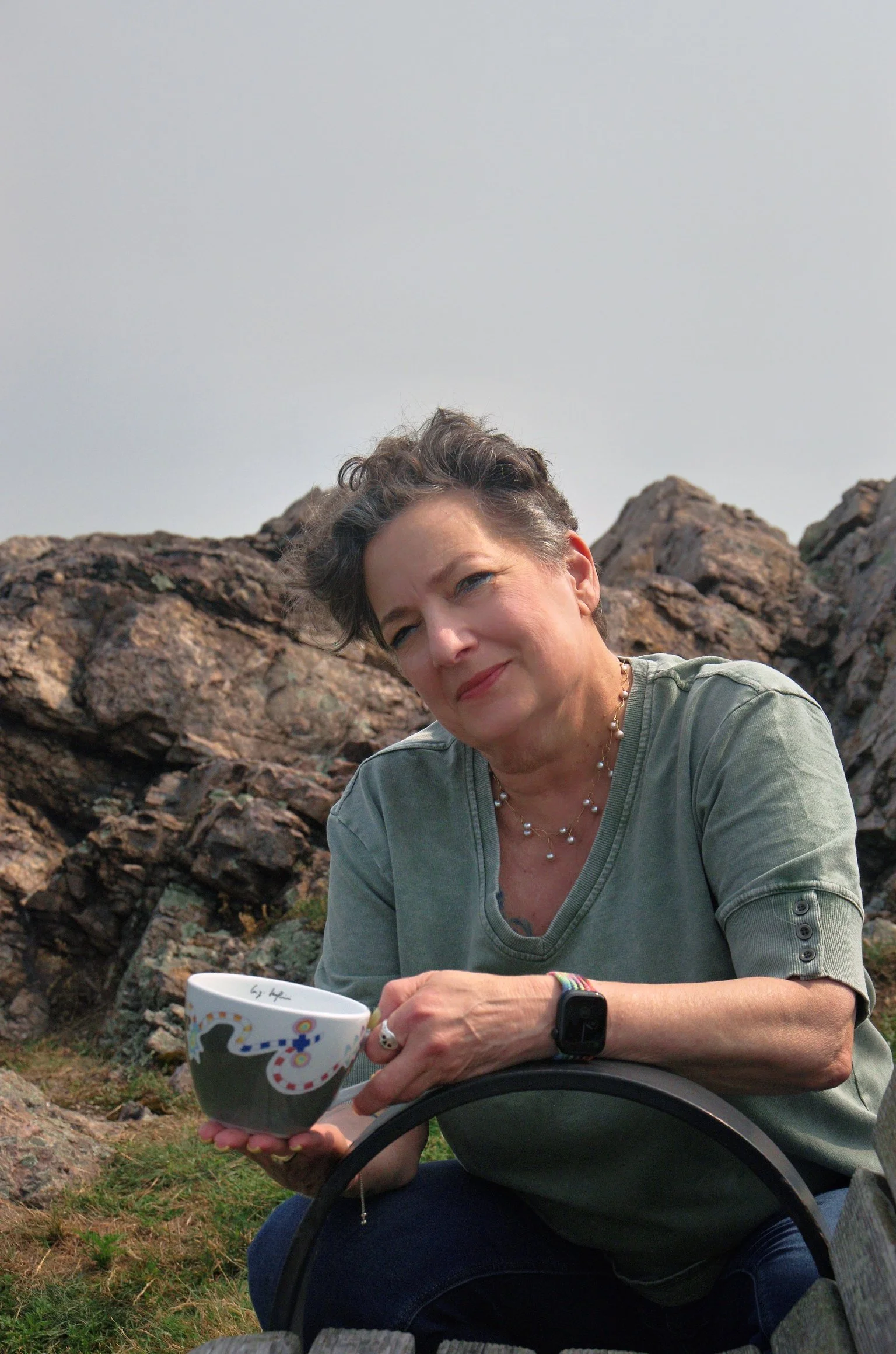 A woman with short curly hair sitting outdoors on a rocky area, holding a colorful mug, wearing a gray shirt and a smartwatch, with a curious expression.