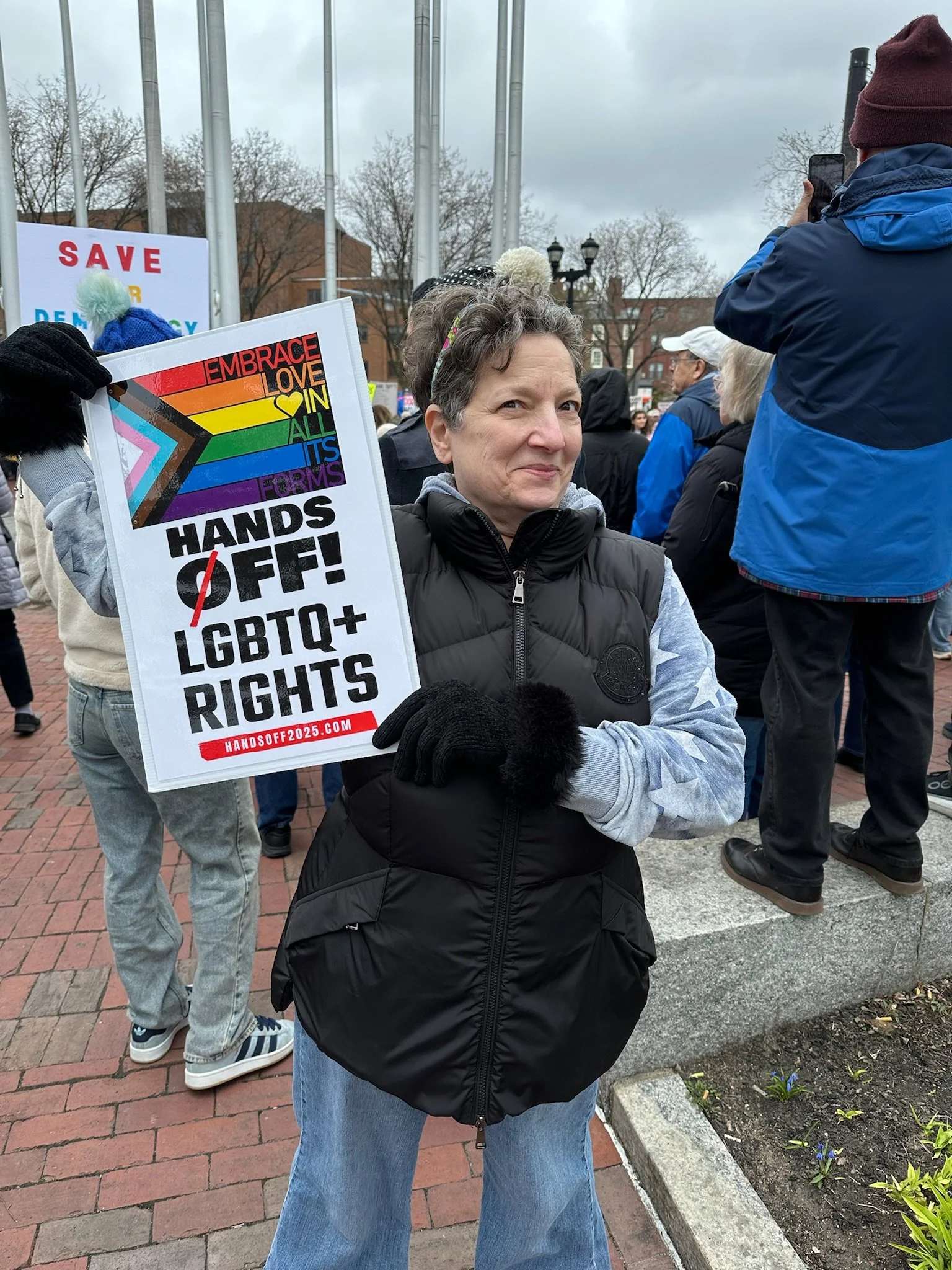 A woman at a protest holding a sign with rainbow pride colors and LGBTQ+ rights messages, wearing a black vest and gray hoodie, with people and trees in the background.