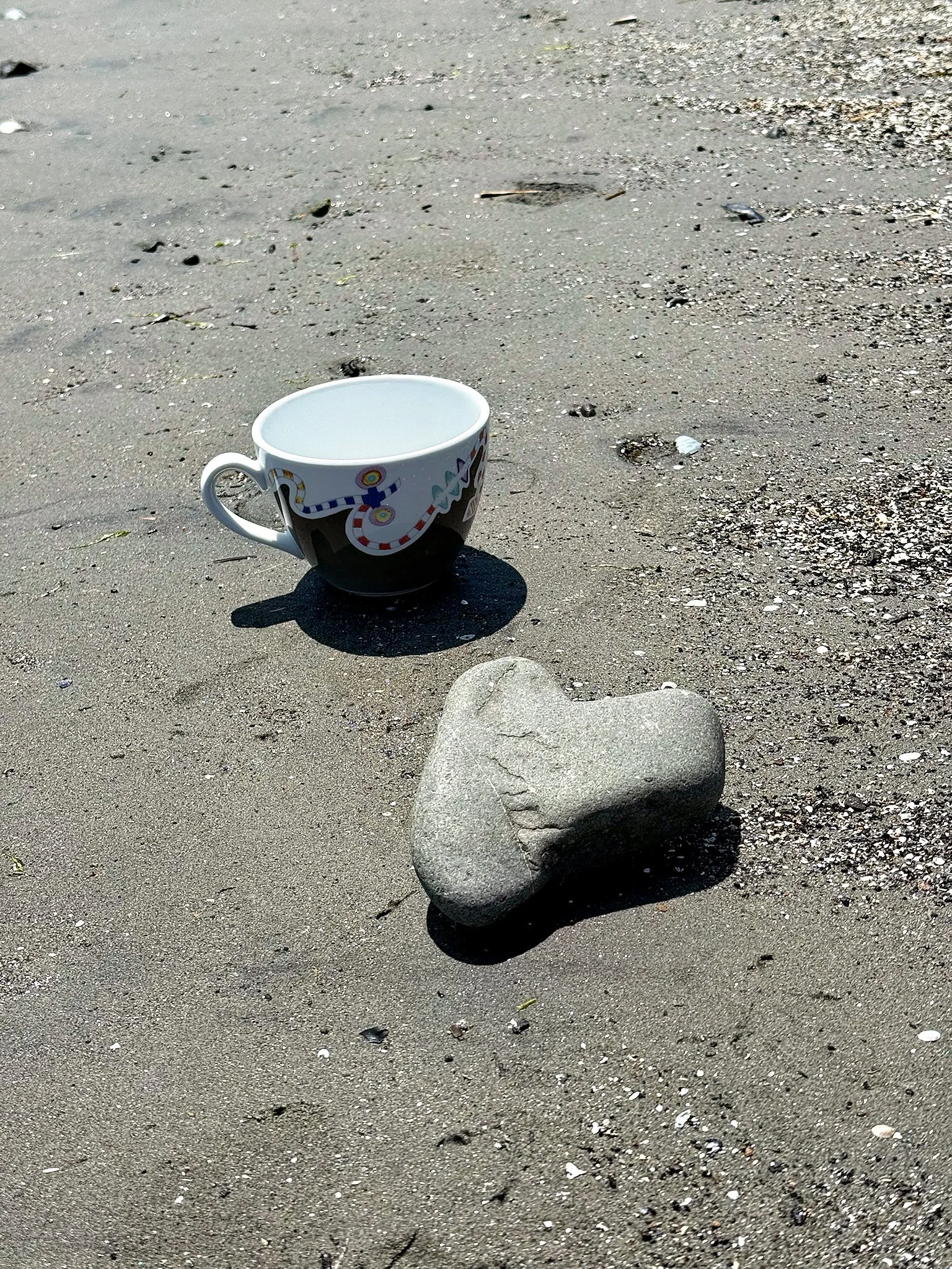 A white ceramic mug with colorful designs sits on the sandy beach, casting a shadow. A large smooth rock is nearby on the sand.